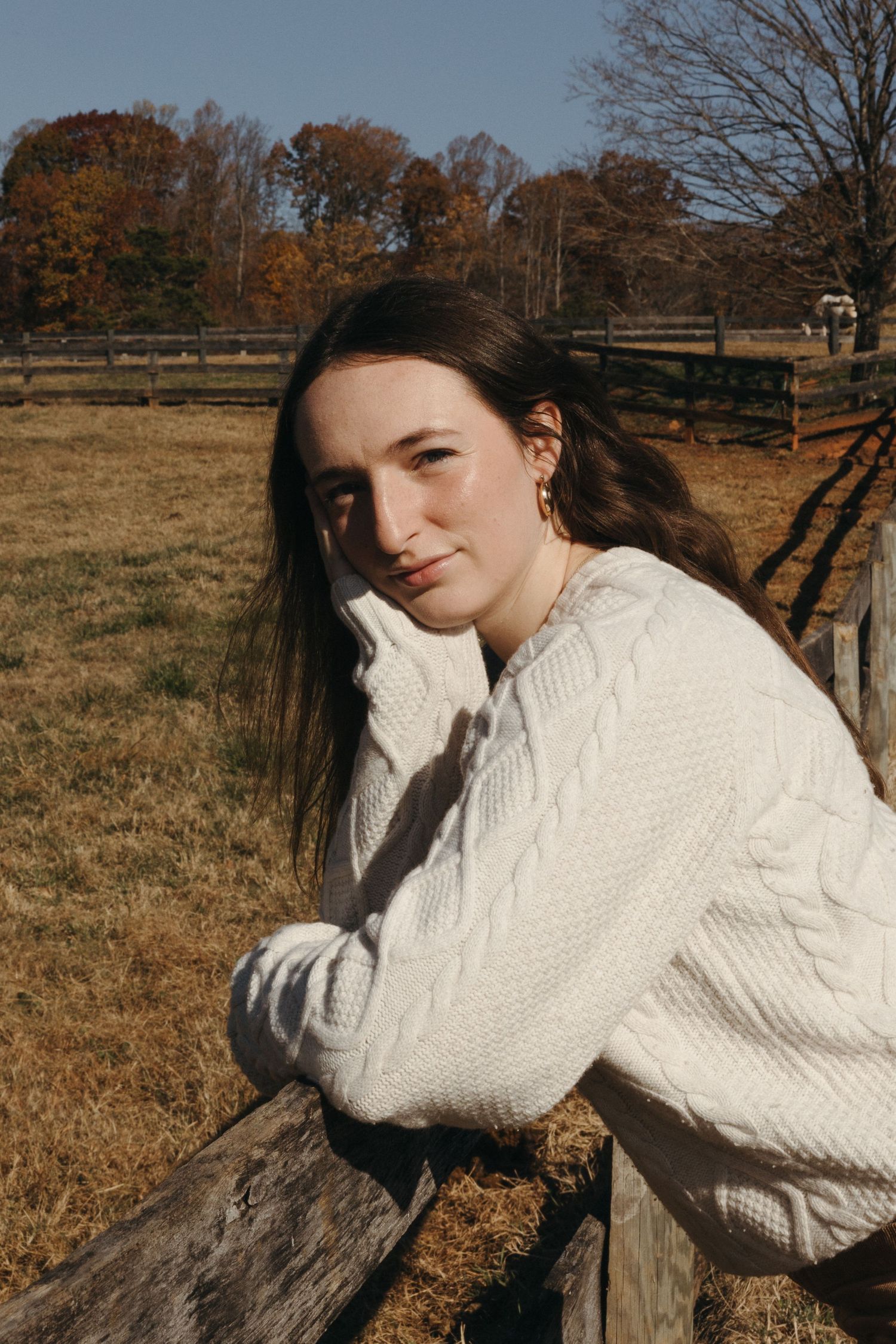 Person in white cable knit sweater poses against rustic wooden fence in autumn field with fall foliage in background.