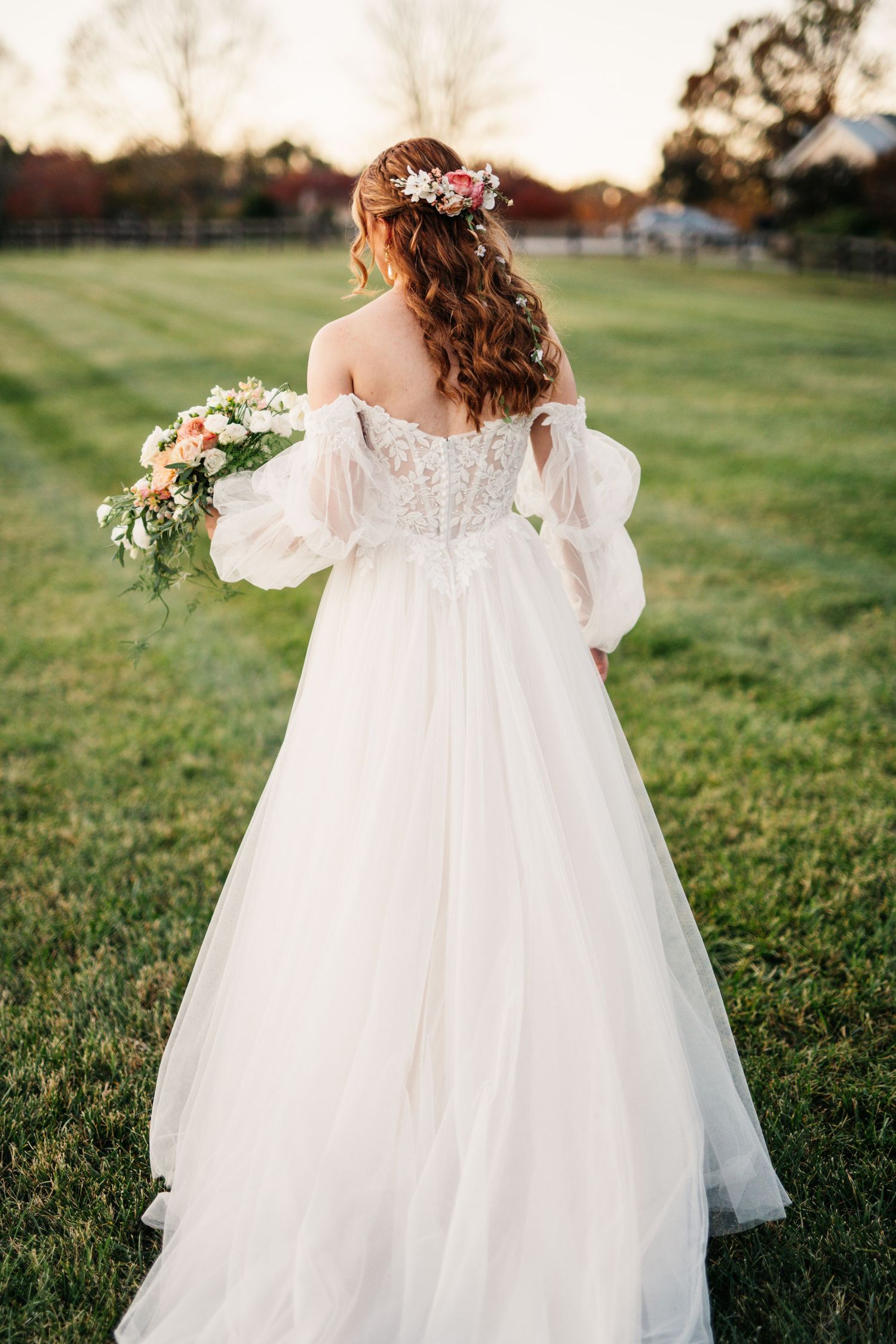 A romantic bridal portrait from behind showing an off-shoulder wedding gown with flowing sleeves and floral crown in a garden.