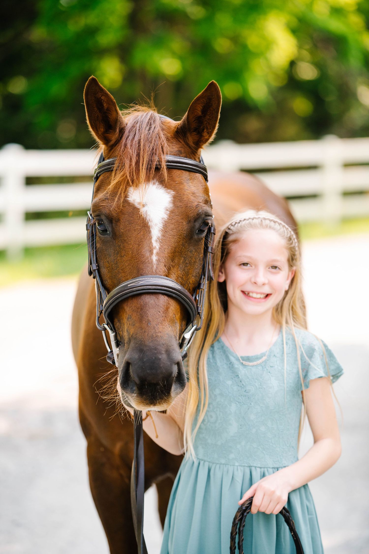 A chestnut horse wearing a black bridle standing next to someone in a blue dress against a white fence background.