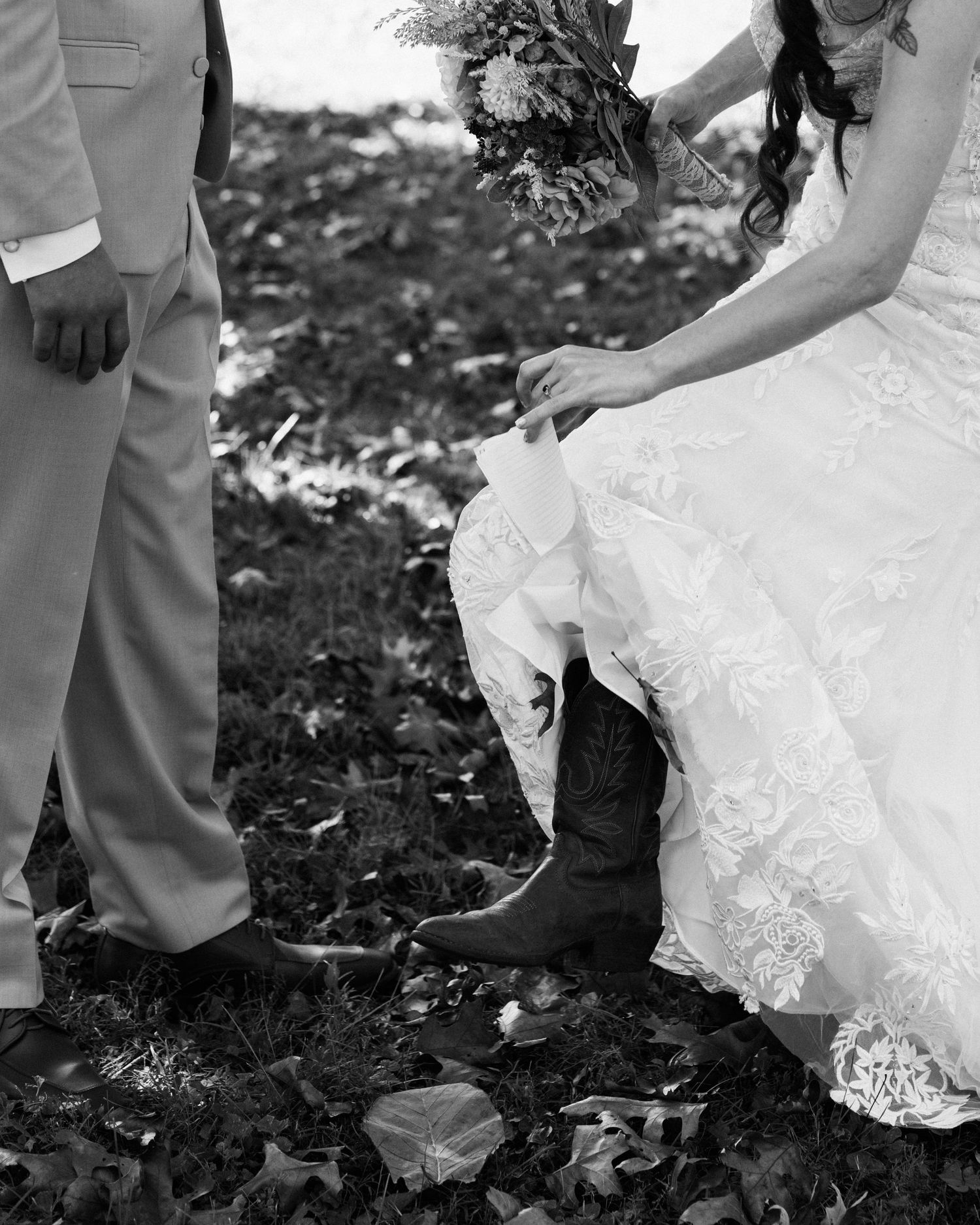 Wedding dress and boots detail shot with groom's pants visible on fallen leaves.