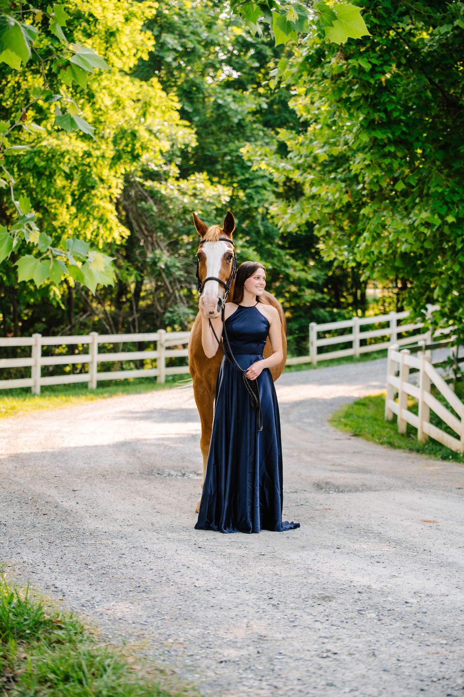 An elegant horse and formal dressed companion pose on a tree-lined gravel path with white fencing.