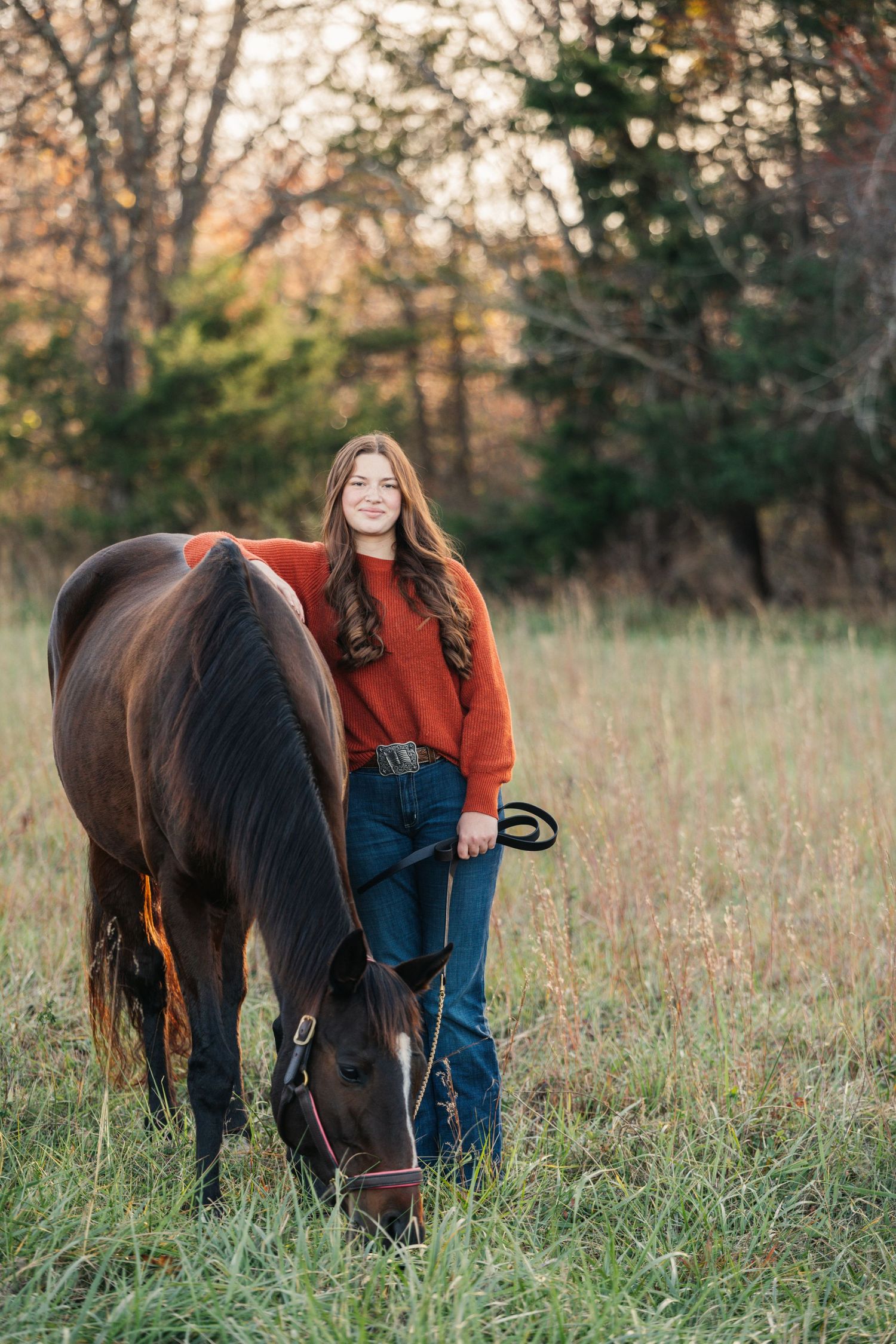 Person in orange sweater stands beside grazing brown horse in autumn field at sunset.