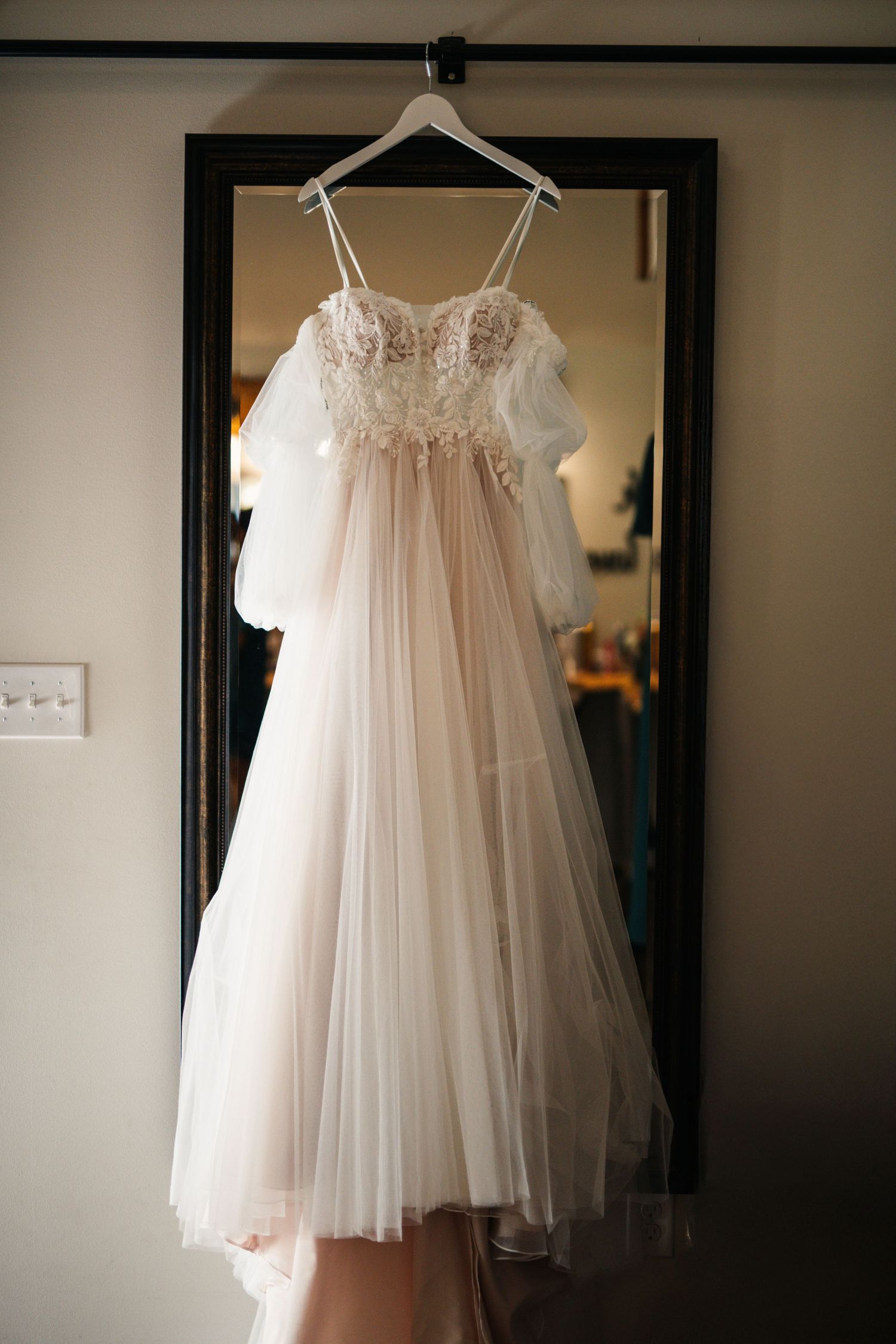 Delicate lace wedding dress with tulle skirt hanging on white hanger against dark doorframe.