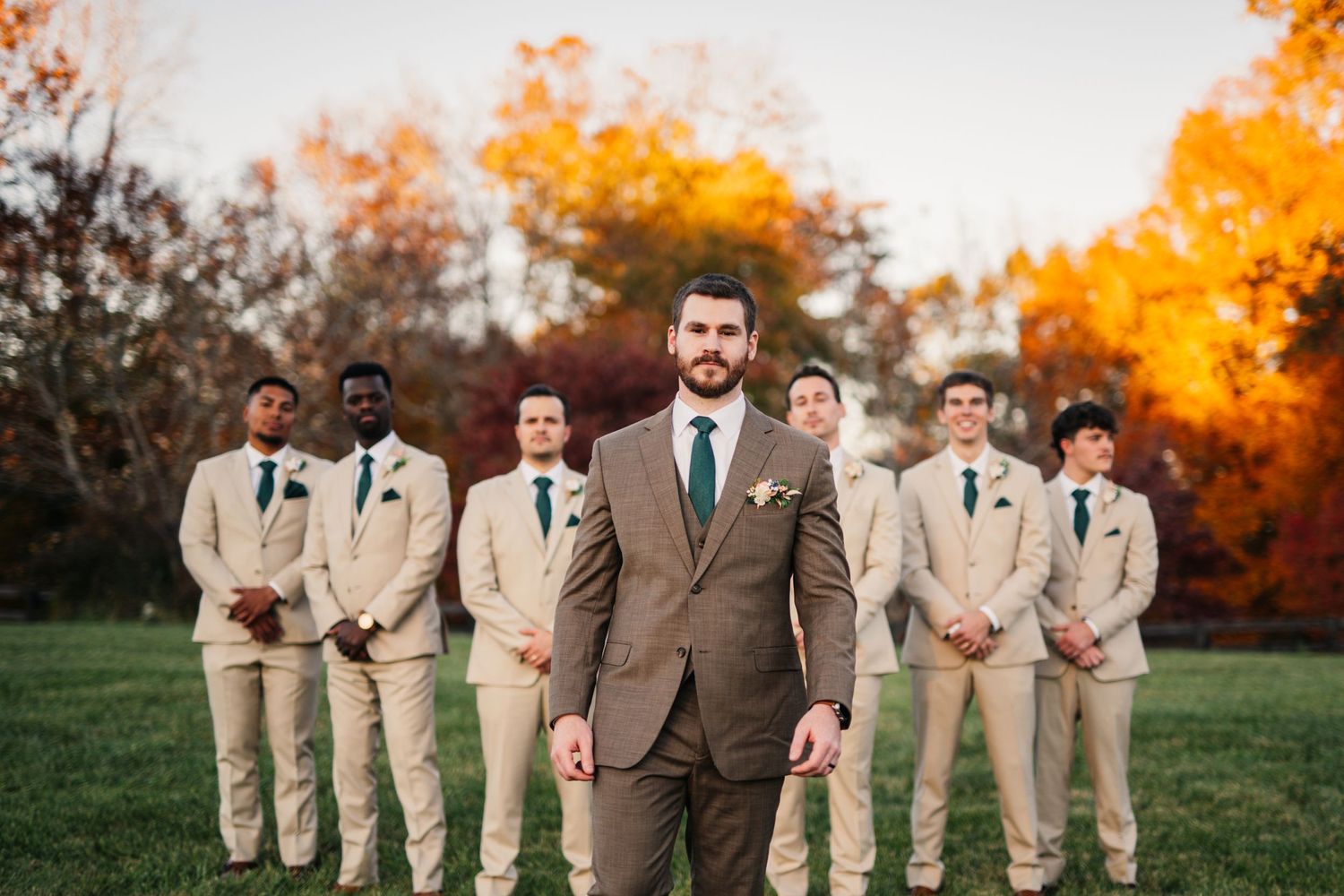 Wedding party in beige suits pose against vibrant autumn foliage at sunset.