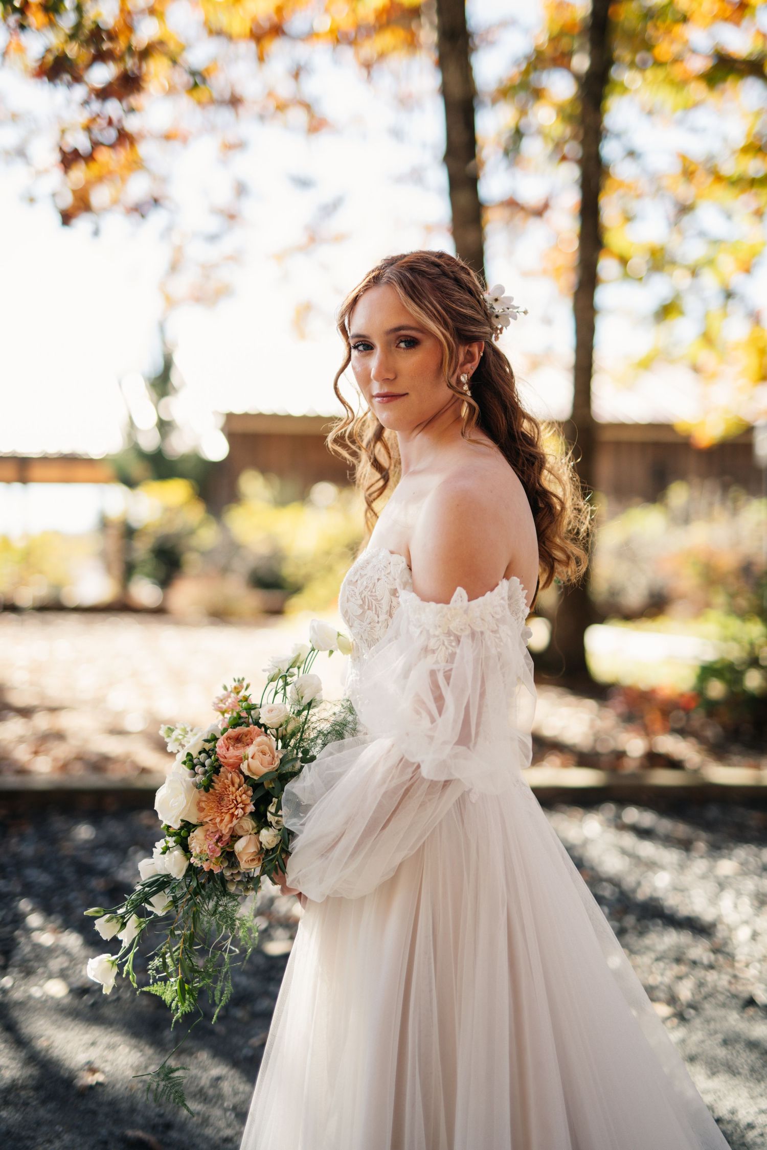 Bride in off-shoulder lace gown holds bouquet in autumn outdoor setting.