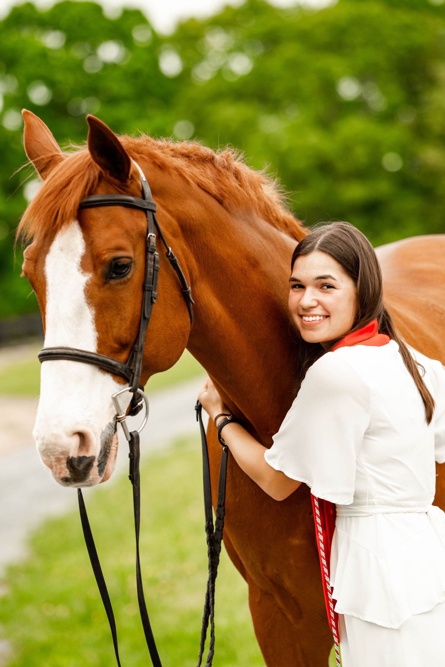 A chestnut horse with white markings poses with someone in graduation regalia outdoors.