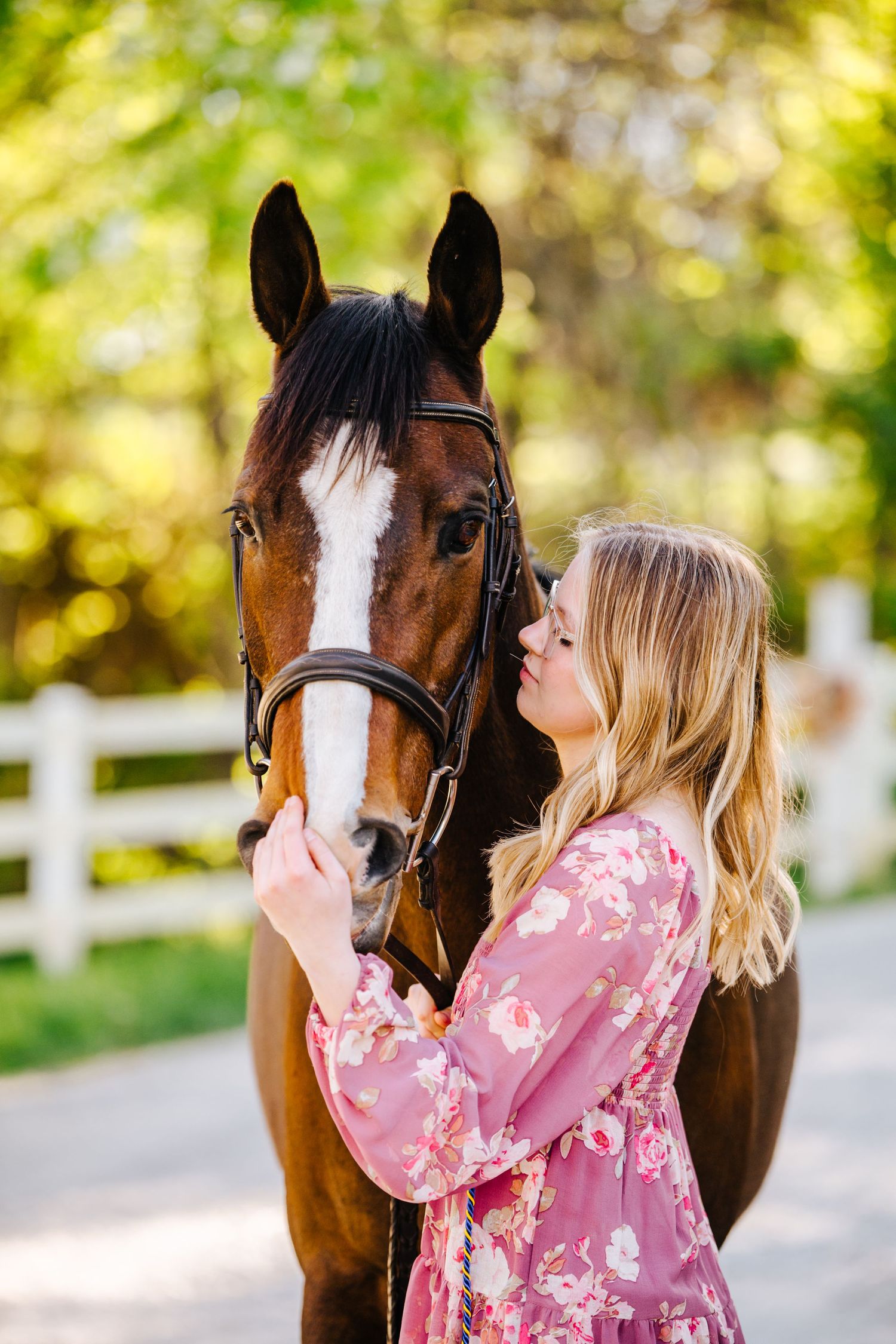 Person in pink floral dress shares affectionate moment with bay horse by white fence.