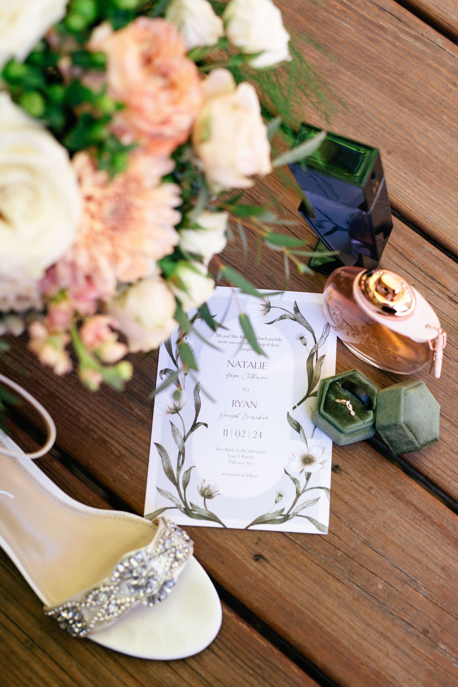 Wedding details with invitation, floral centerpiece and bridal shoe on wooden table.