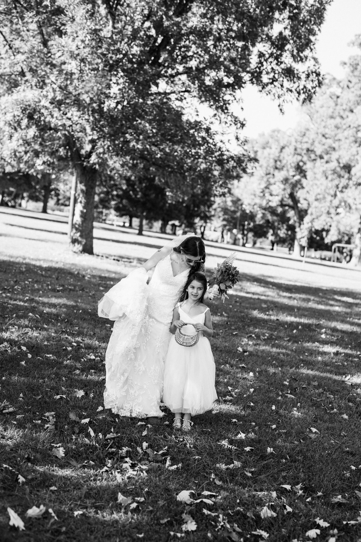 A tender moment between a bride in white and a small flower girl standing on grass under trees at an outdoor wedding.