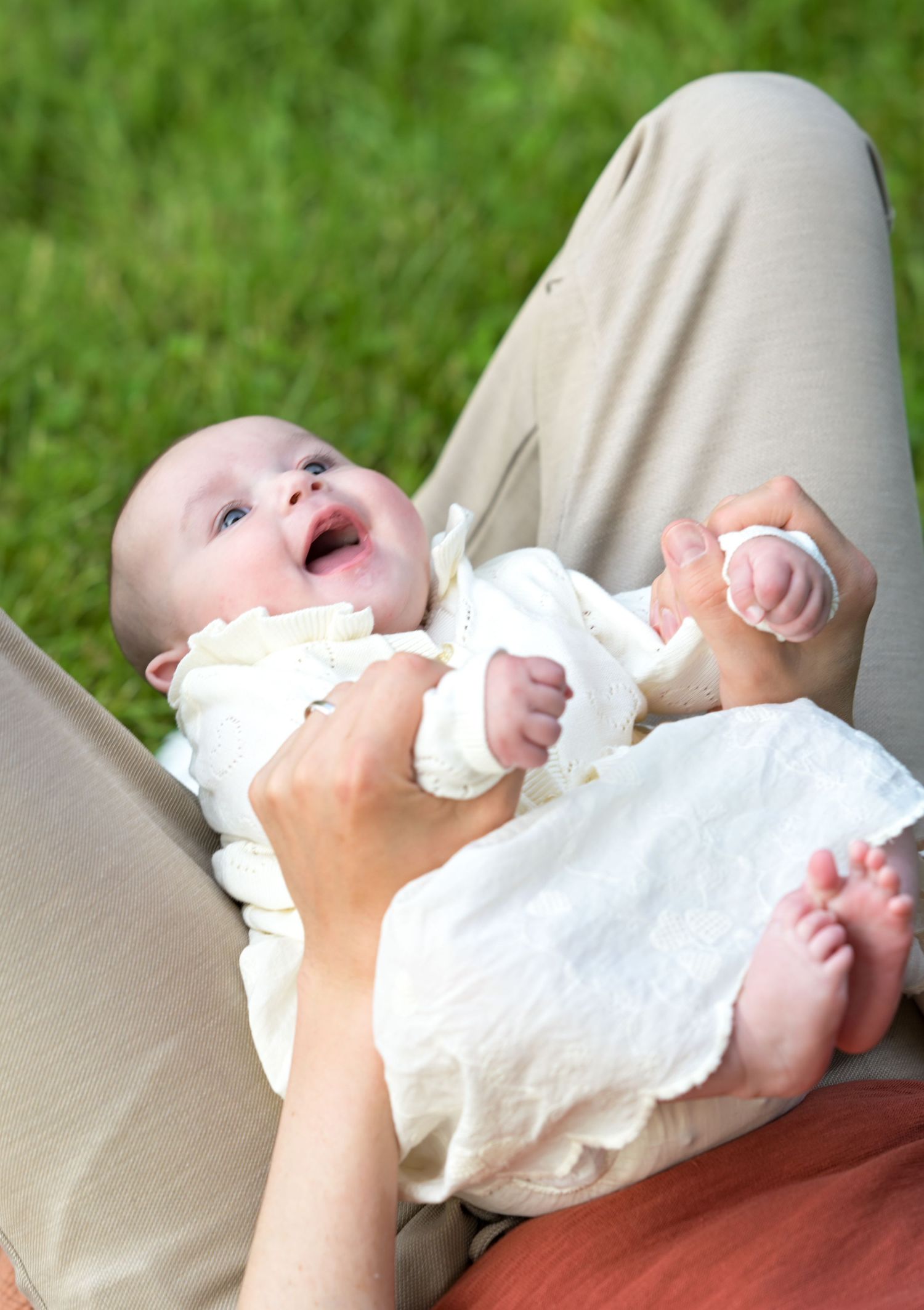 A joyful baby in a white dress lying on someone's lap outdoors on green grass.