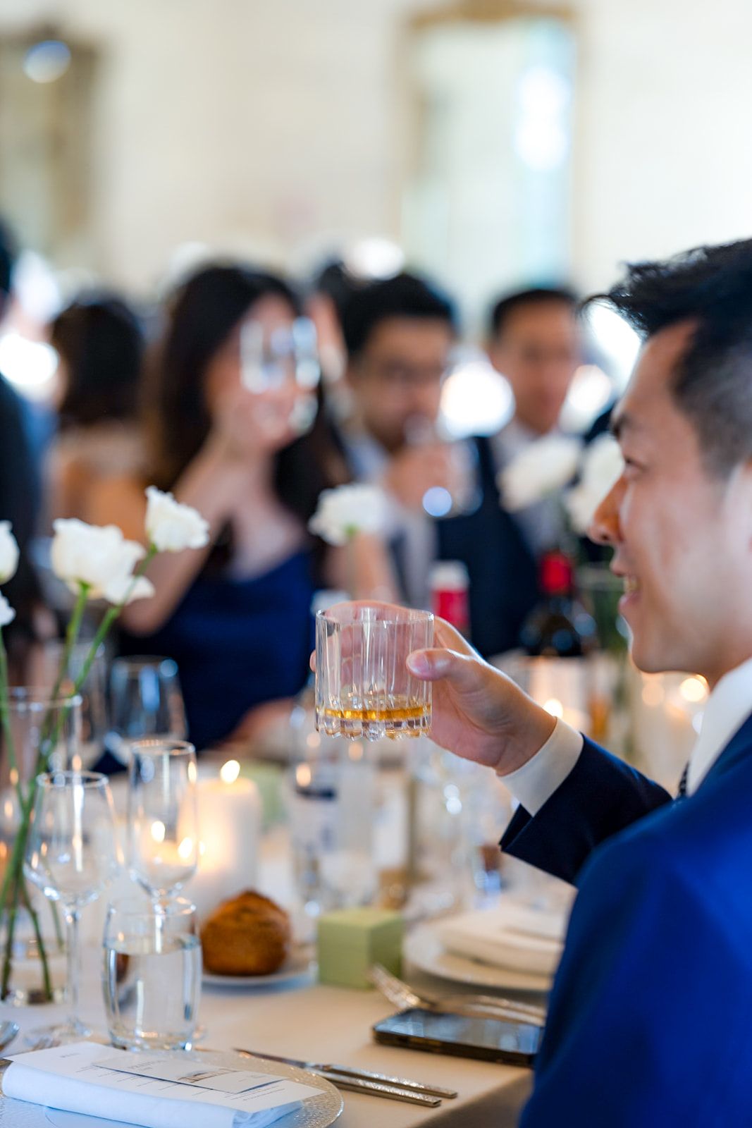 Guest in blue suit enjoying drink at elegant reception with floral centerpieces.