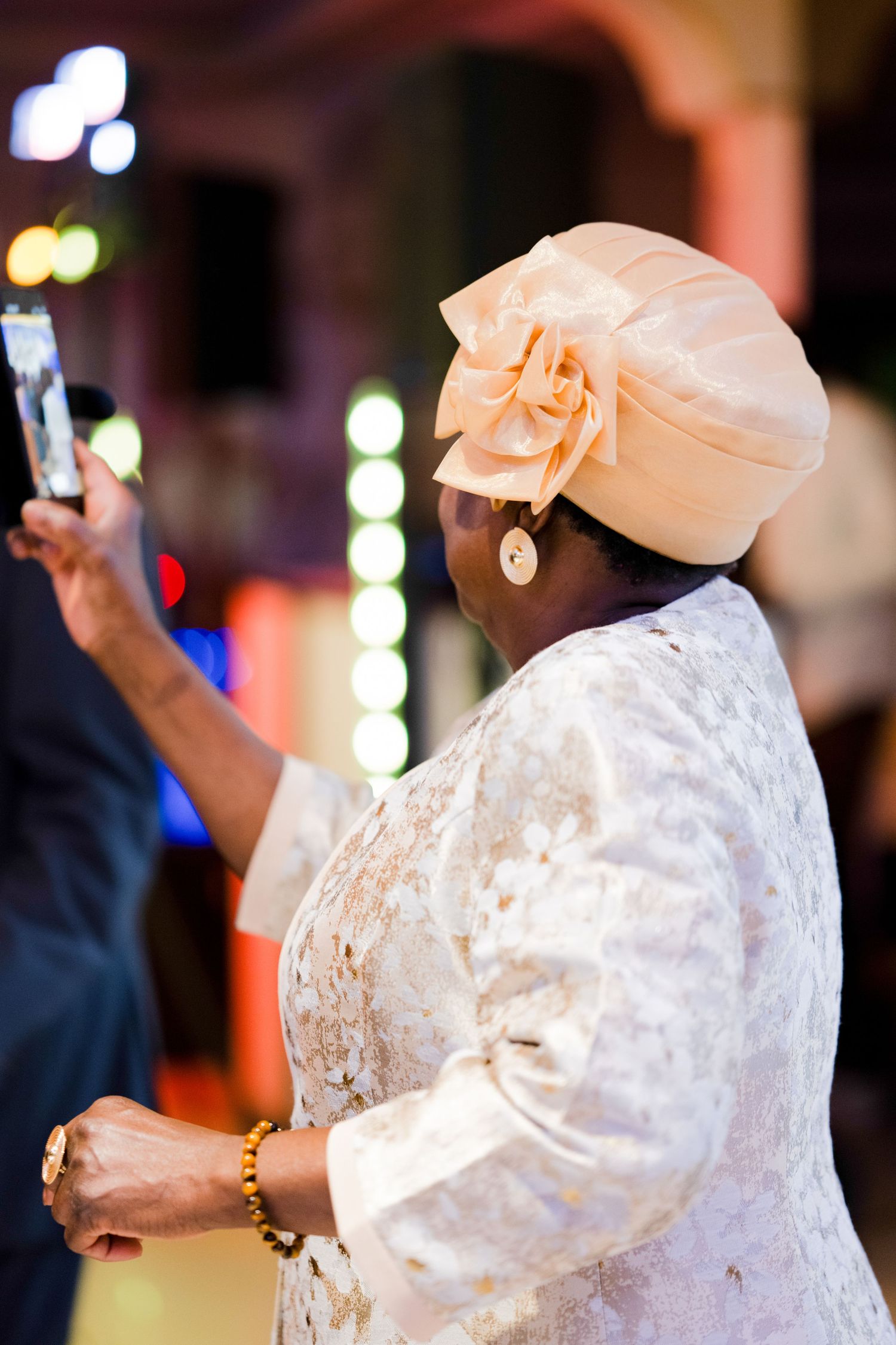 Wedding guest in elegant white lace dress and peach headpiece holds up phone at celebration.