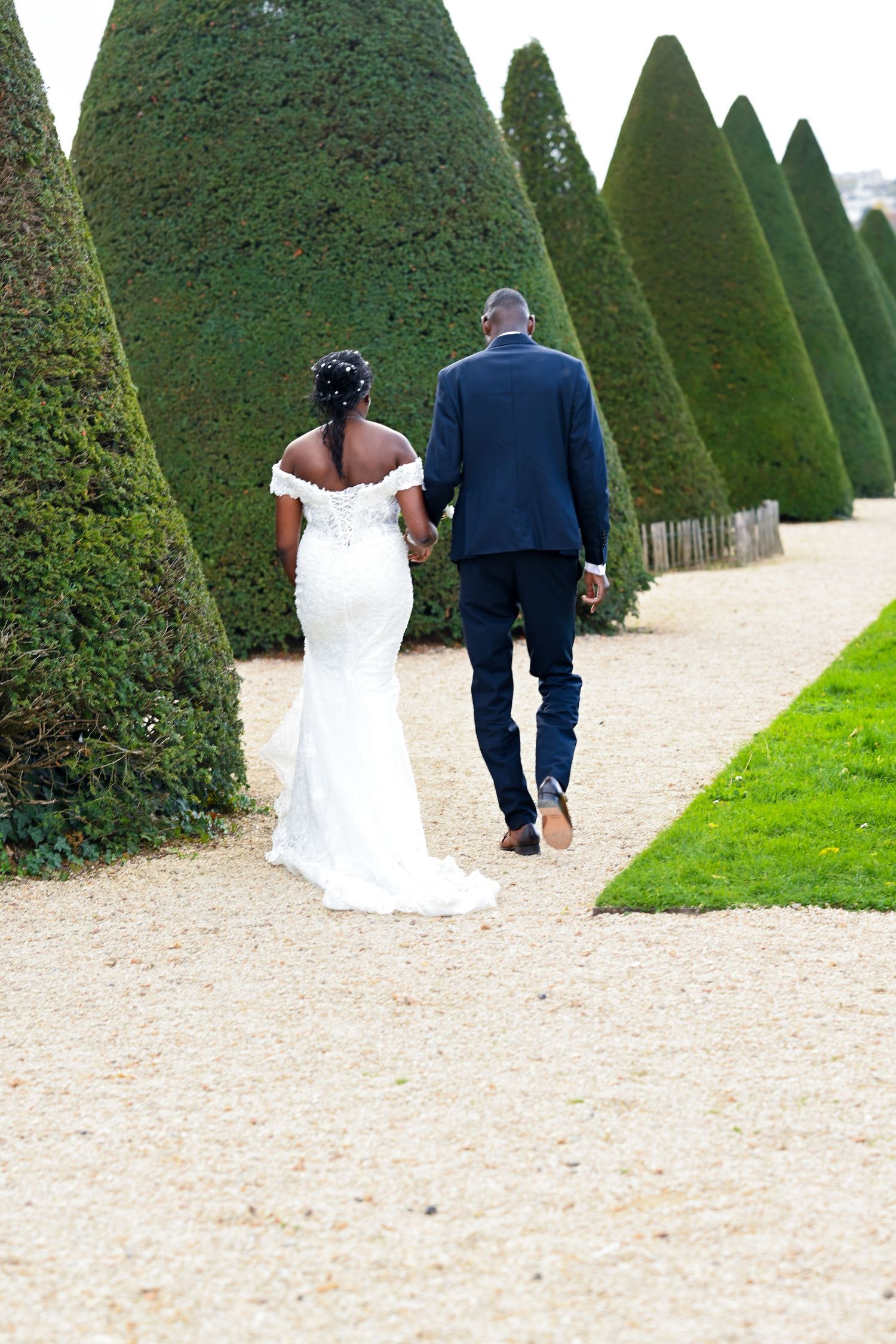 Newlyweds walk hand in hand down a formal garden path lined with perfectly trimmed topiary trees.