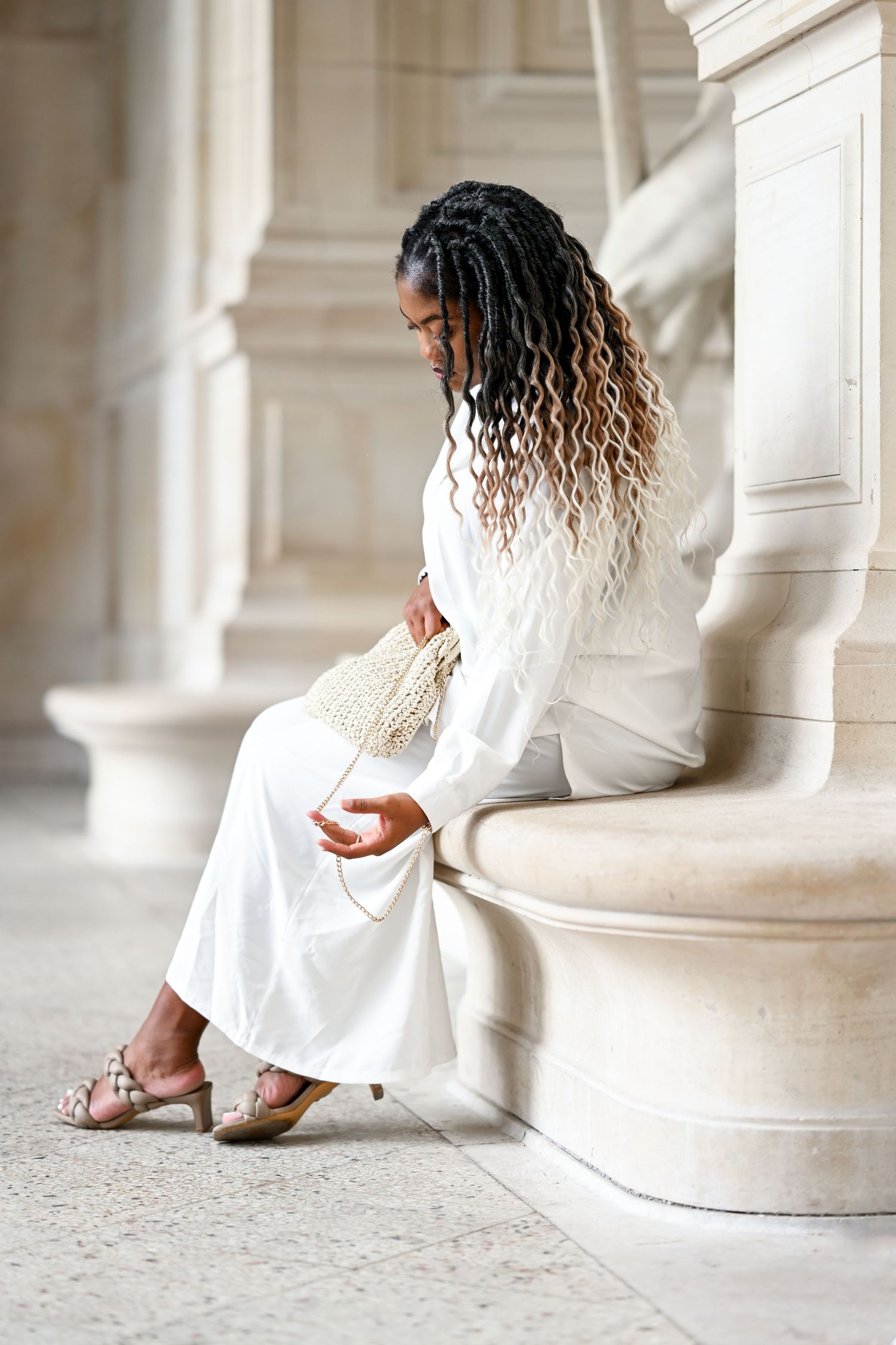 Side profile of model in white attire seated on classical marble column with ornate details.