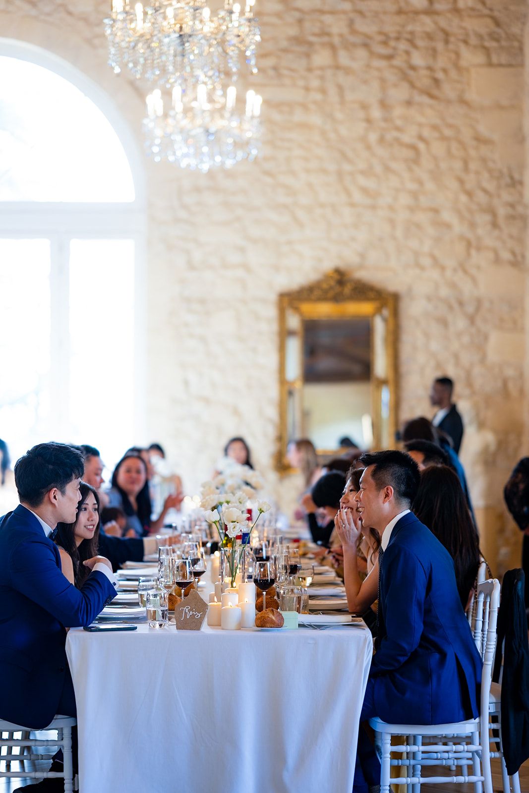 Elegant wedding reception with guests seated at long tables in a bright stone-walled room.