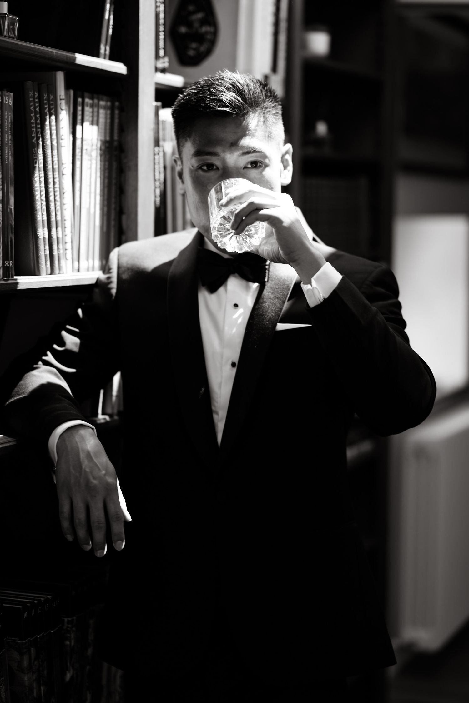Classic black and white photo showing a man in tuxedo drinking from a glass near bookshelves.