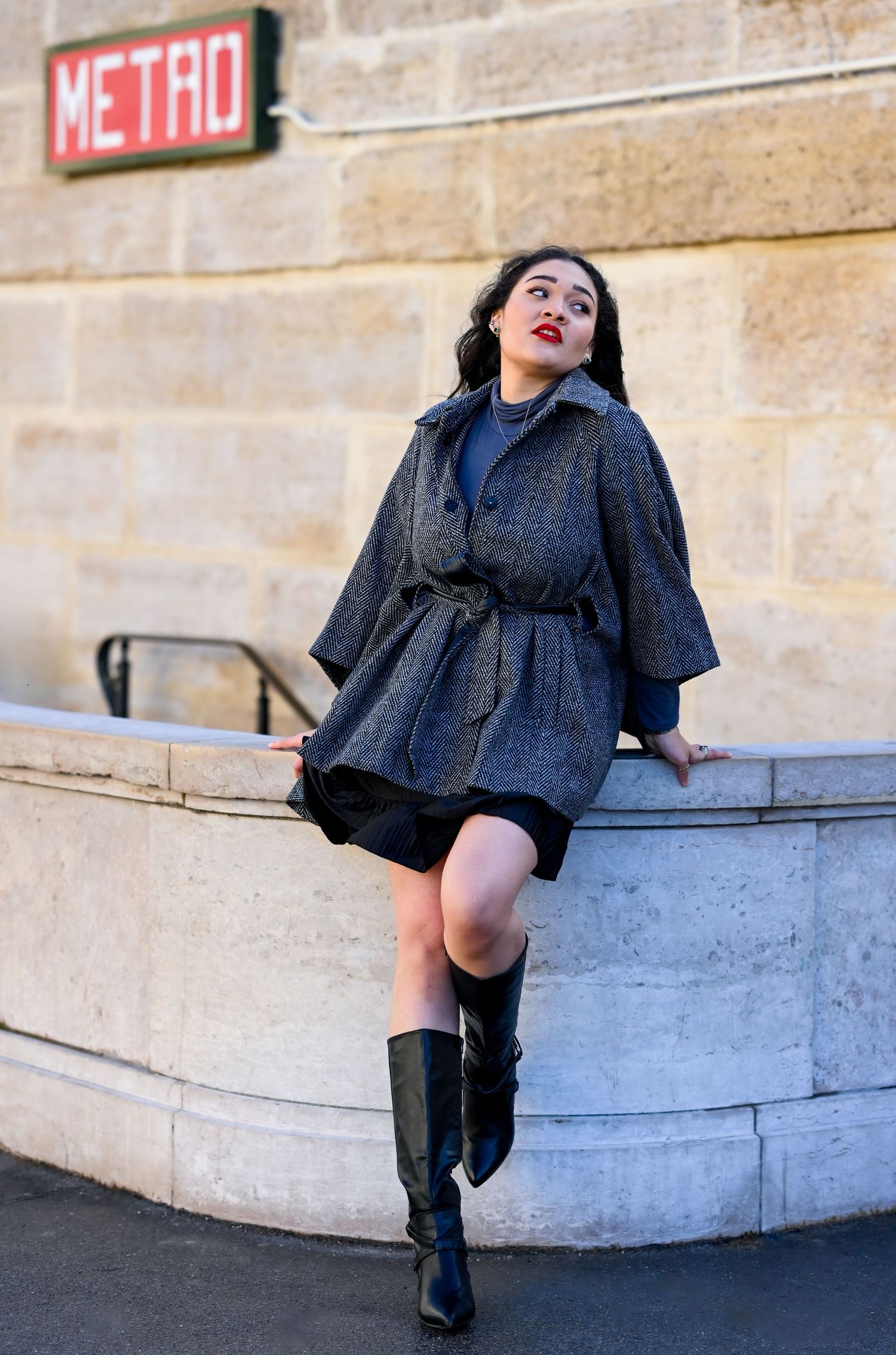 Fashion model in grey coat and boots poses playfully by Metro sign in Paris.