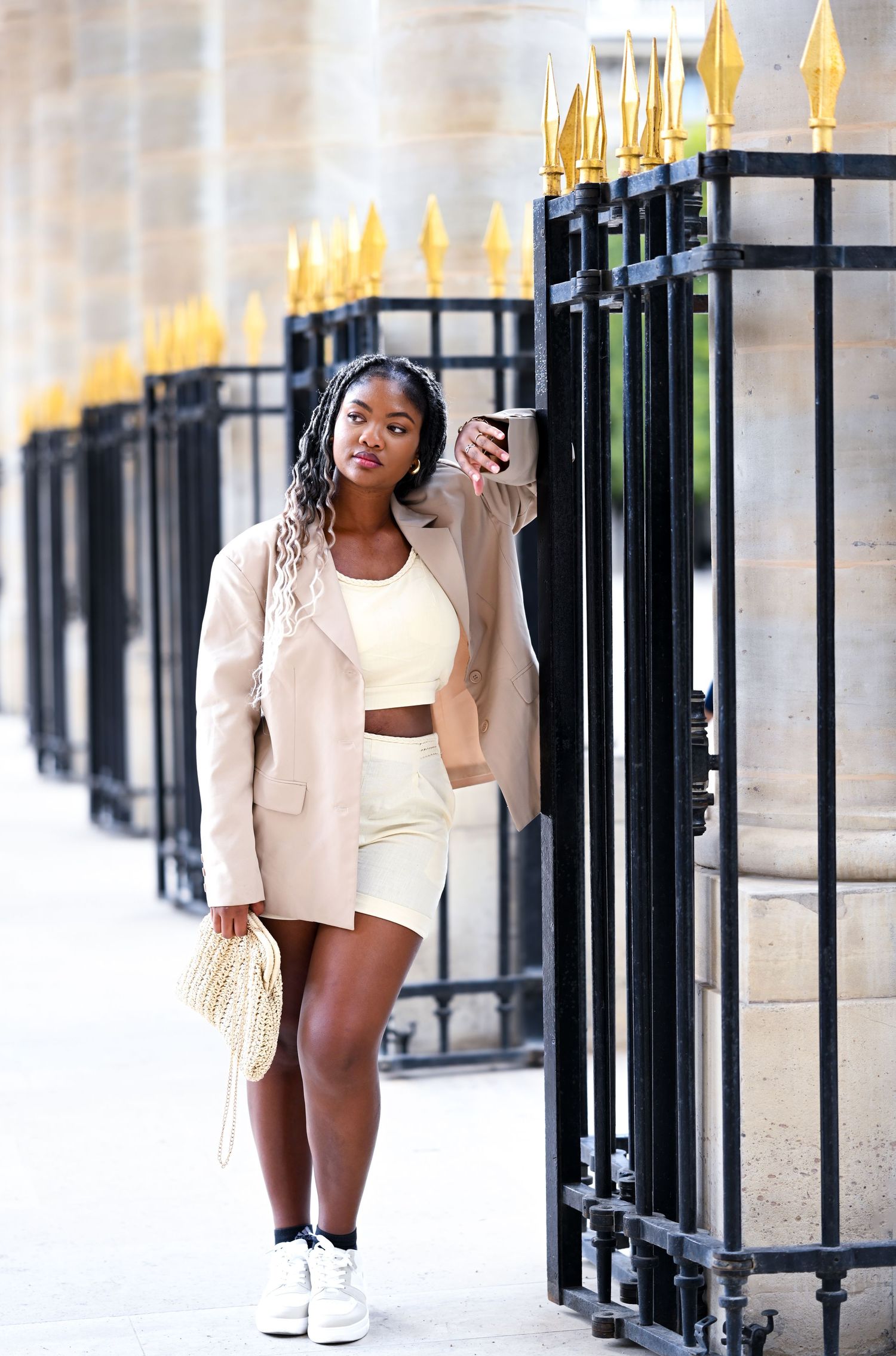 Casual chic outfit with cream blazer and woven handbag against decorative gates.