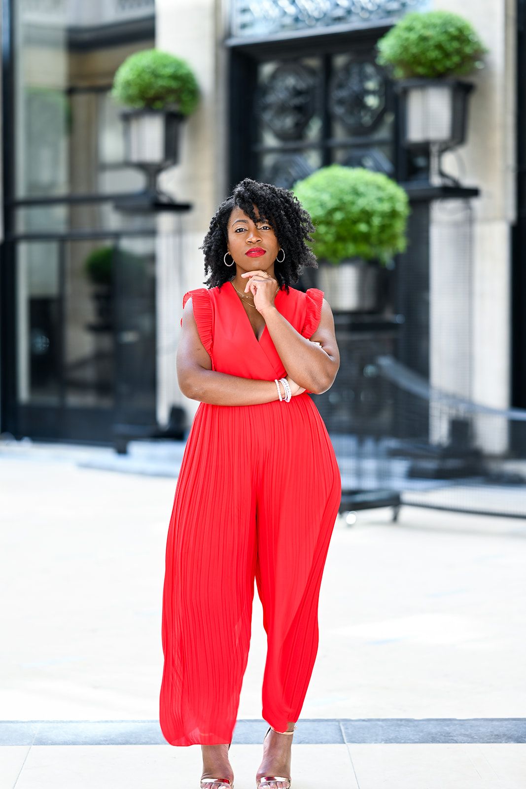 A vibrant red pleated jumpsuit styled with hanging planters and architectural details in the background.