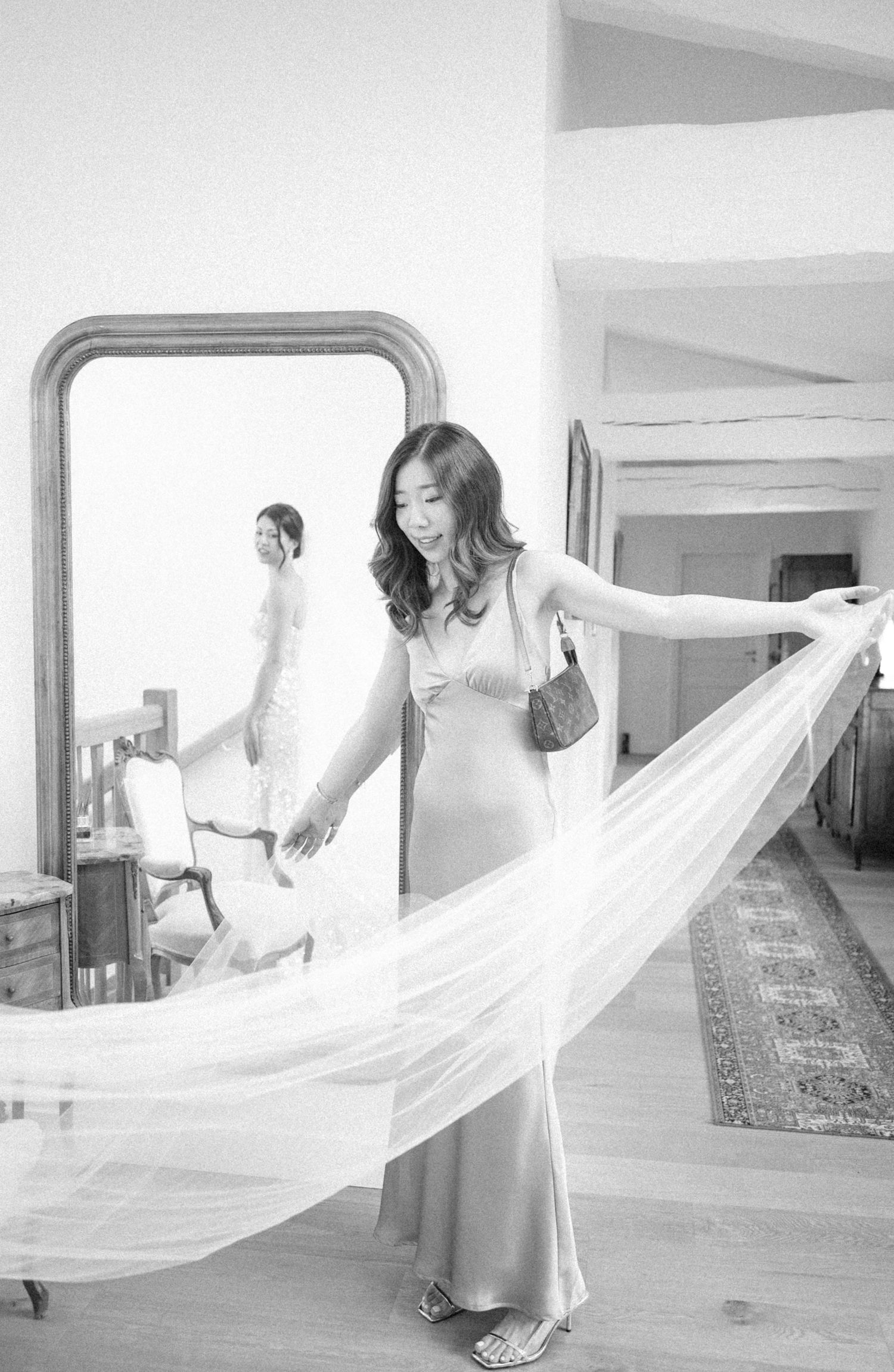 Black and white photo of a woman in a satin gown holding a flowing veil, reflected in a large ornate mirror.