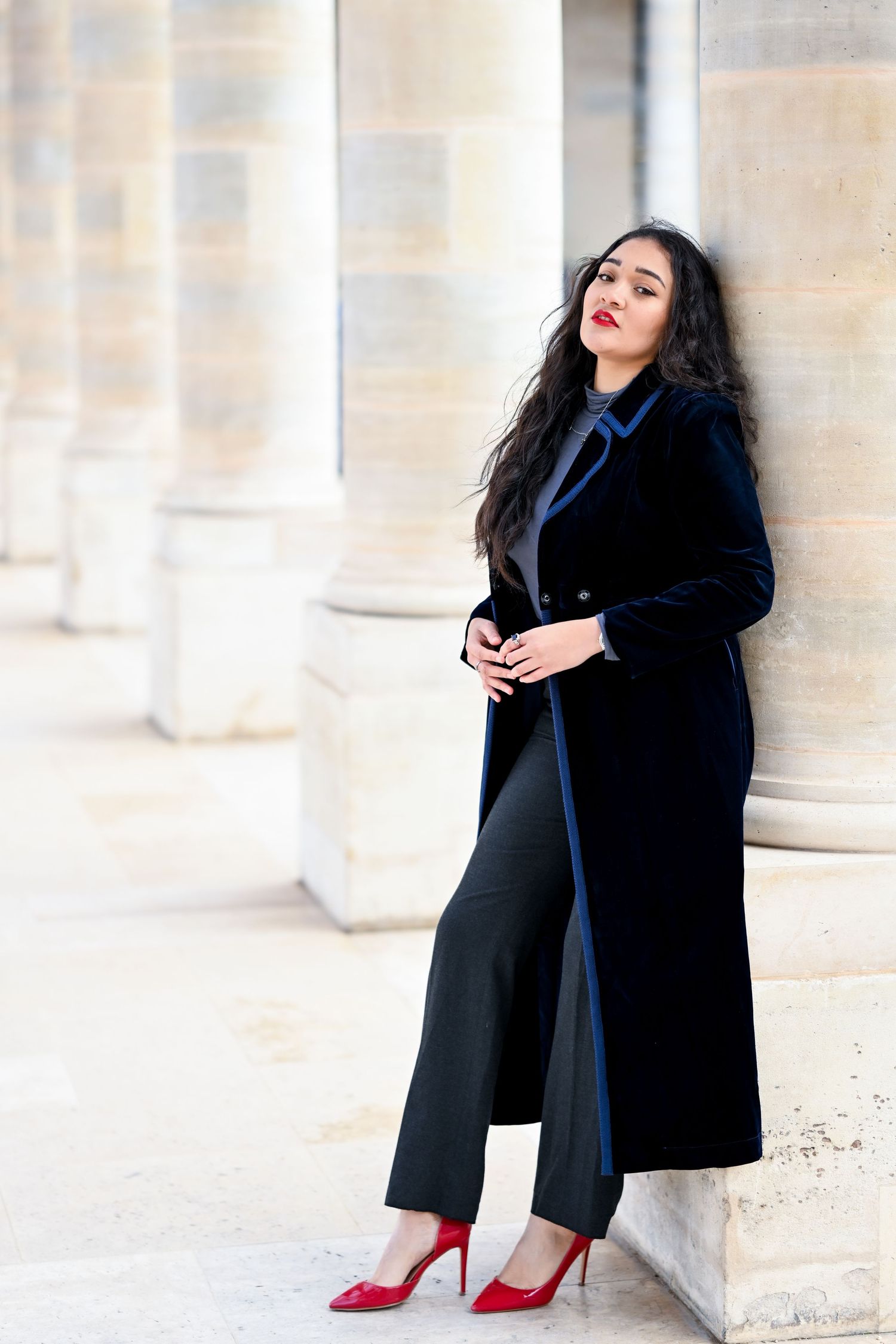 Elegant woman in long black coat and red heels poses against classical stone columns.