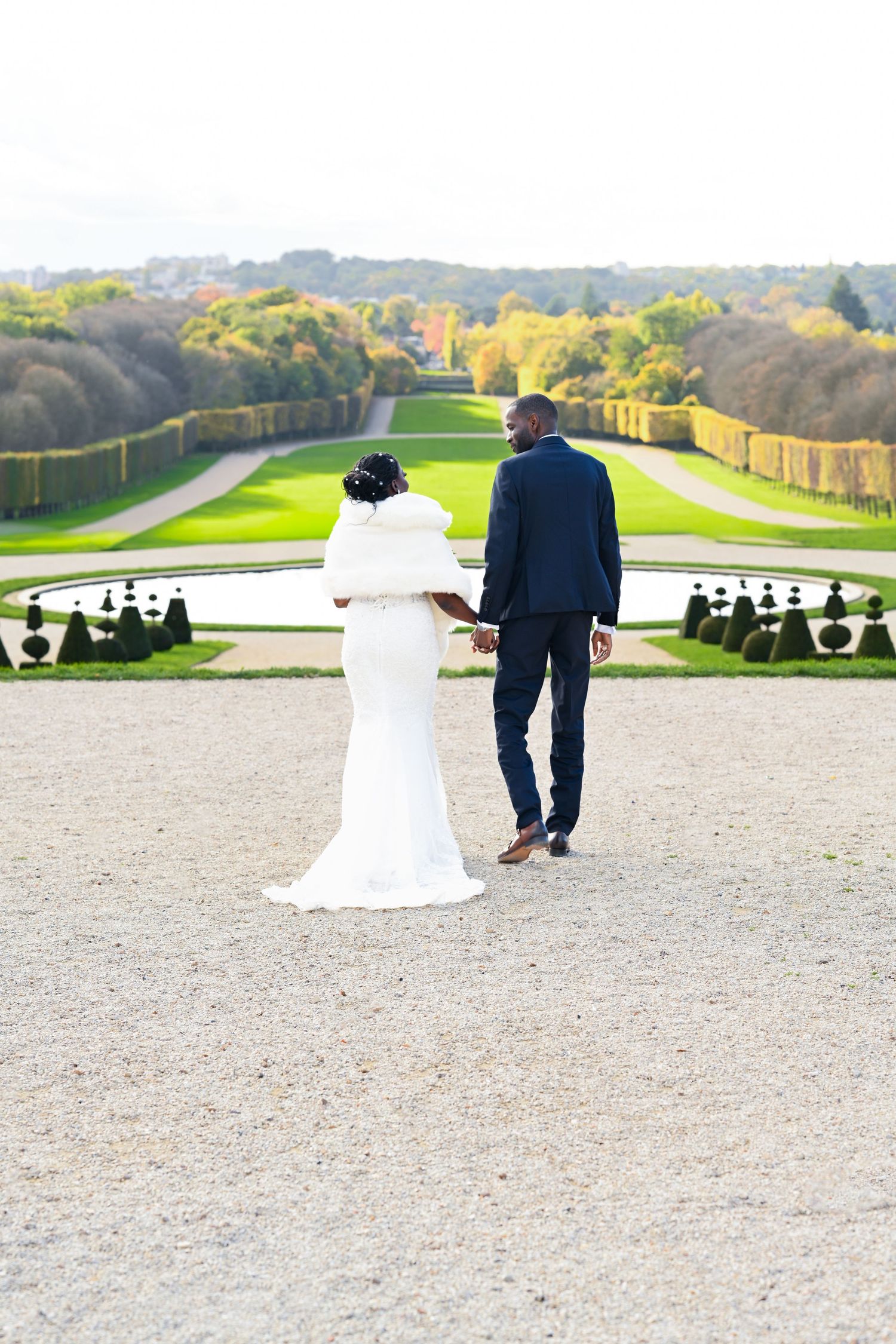 Wedding couple walks away on gravel path with sculptured garden and rolling hills in background.