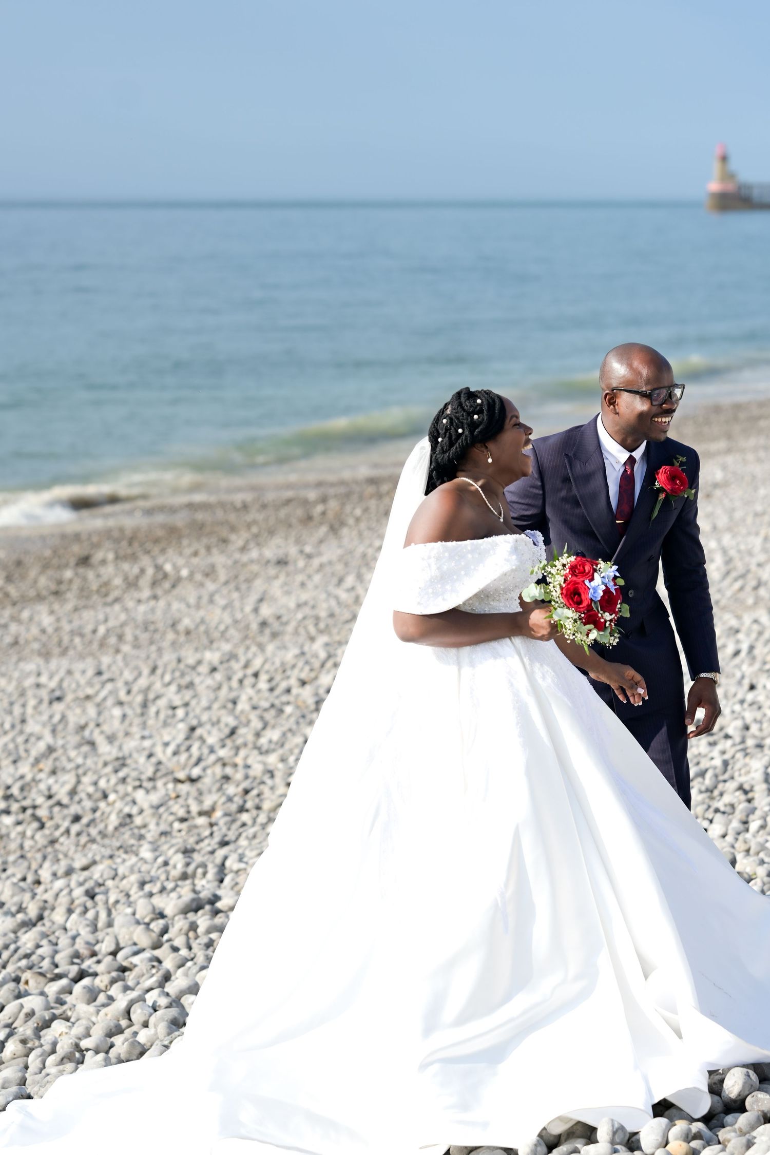 A wedding couple shares a moment on a beach with a lighthouse visible in the distance.
