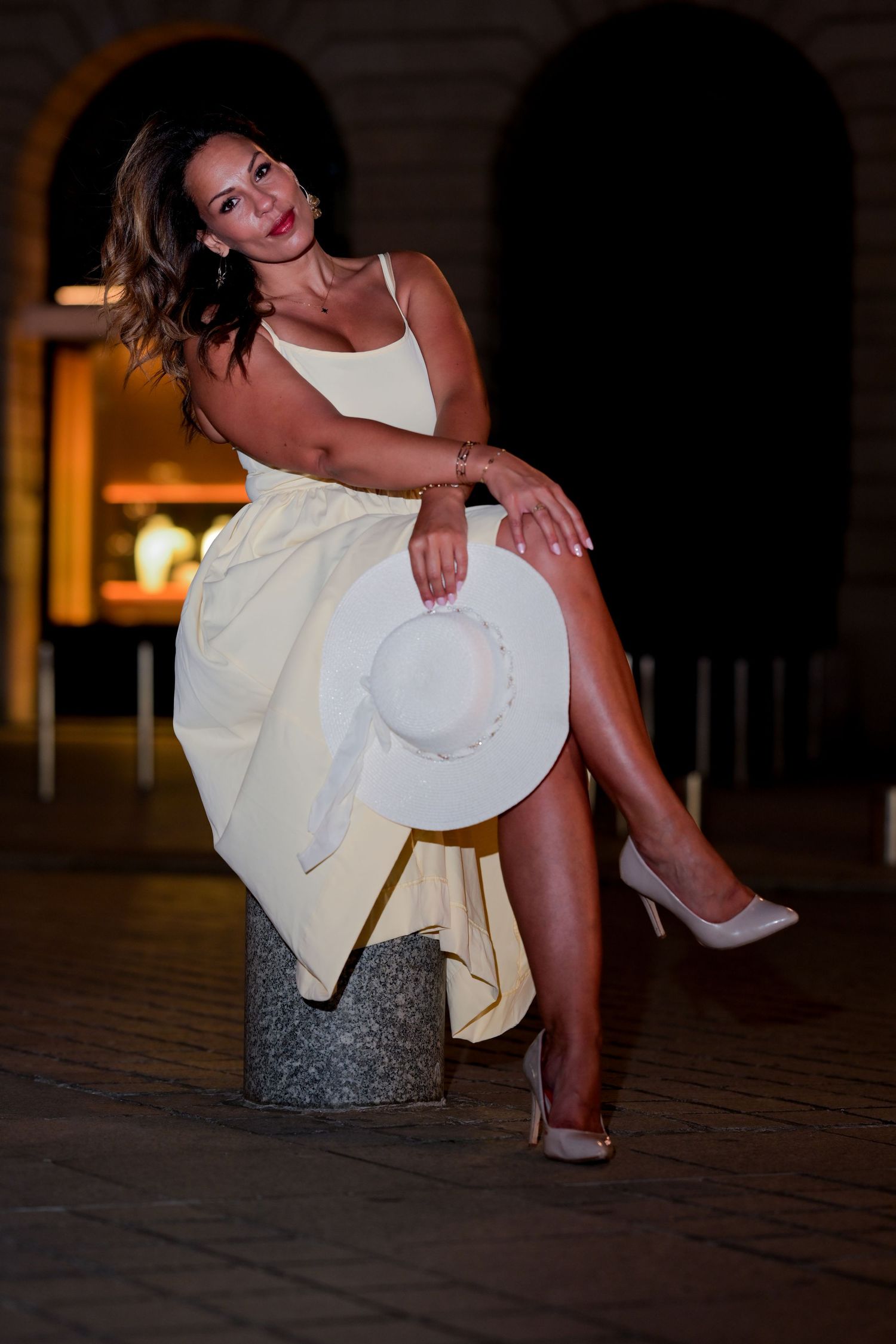 Evening fashion shoot featuring cream dress and white sun hat against dark archways.