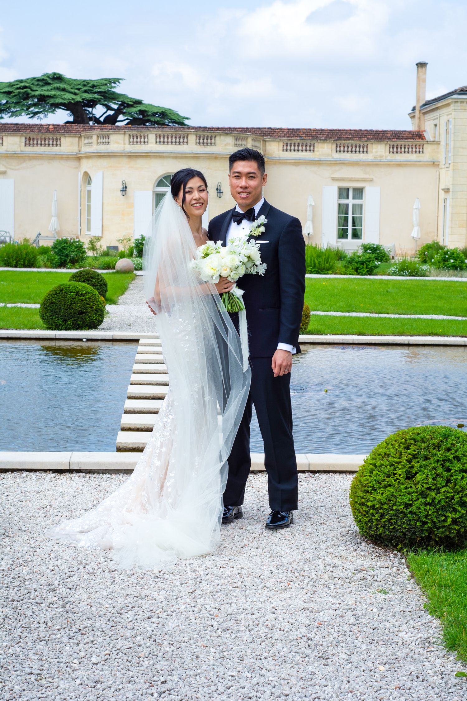 Newlyweds posing by reflecting pool at elegant French chateau venue.