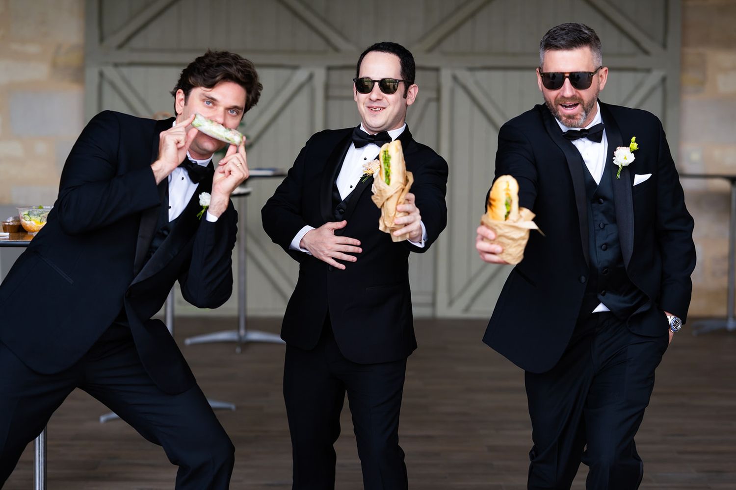 Groomsmen in black tuxedos pose playfully with drinks at a wedding reception.