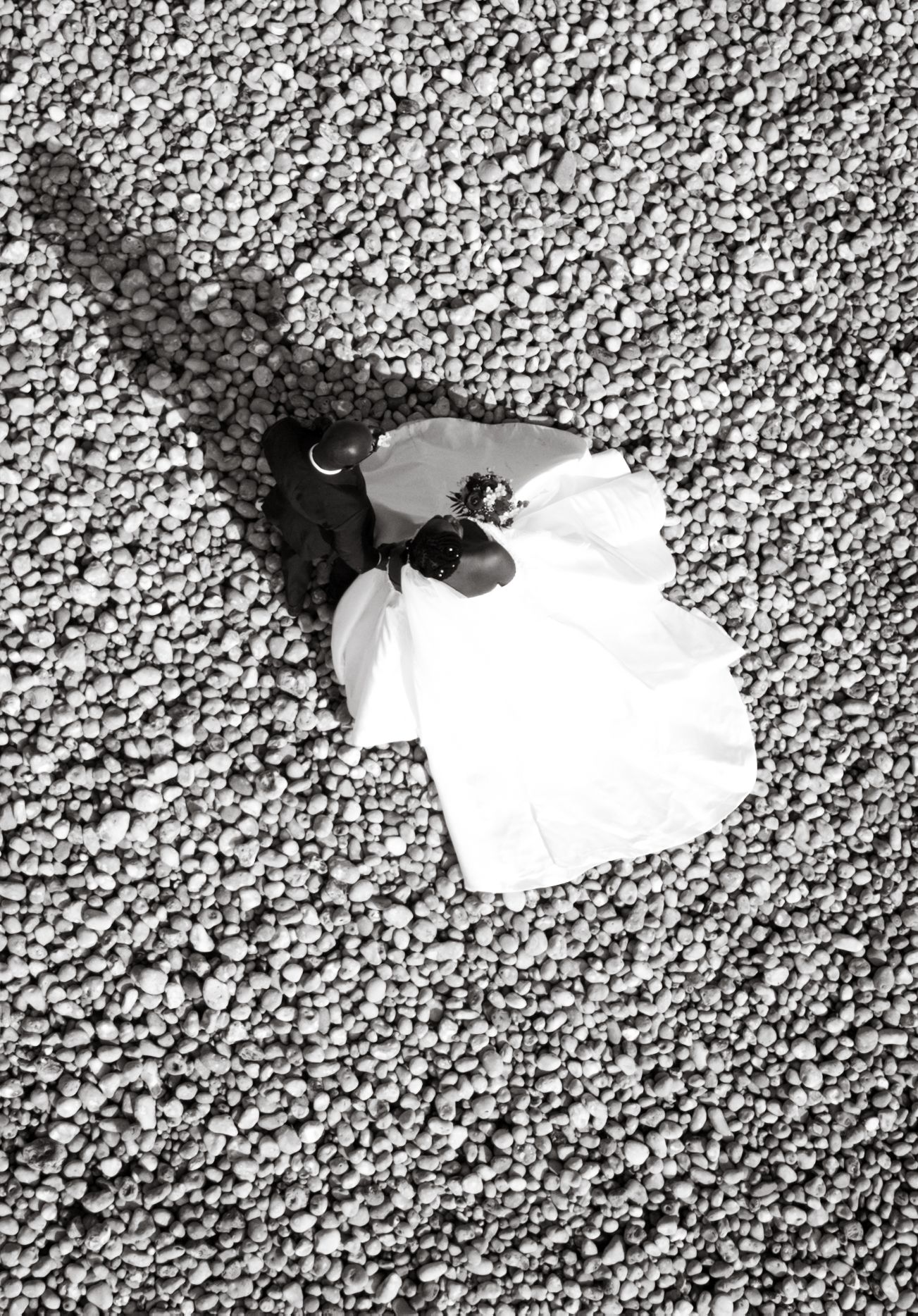 Aerial black and white view of a bride in white dress lying on a pebble beach.