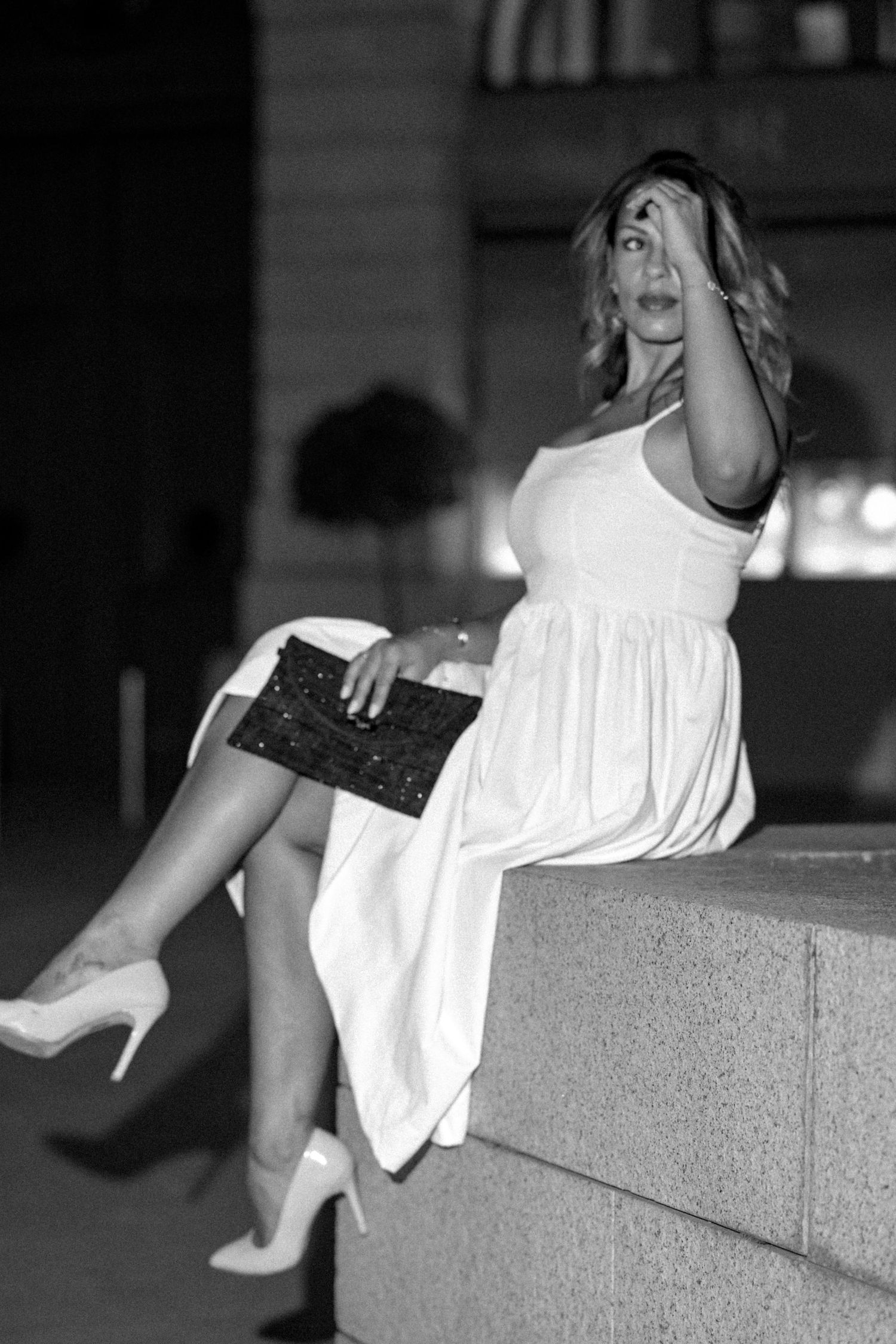 Black and white portrait of woman in white dress posing on stone ledge with clutch.