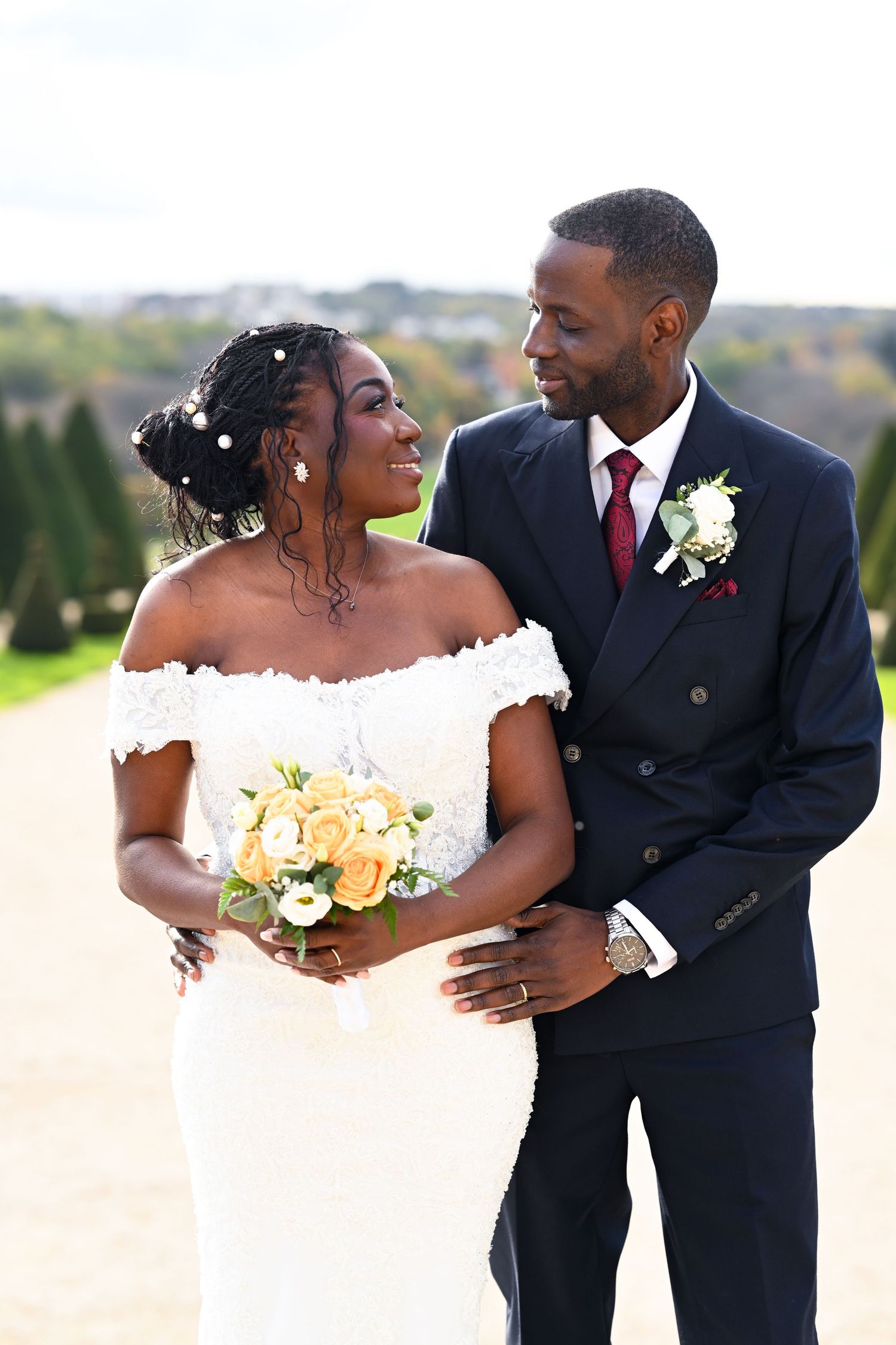 Romantic wedding couple embraces while holding a peach and white bouquet in a formal garden setting.