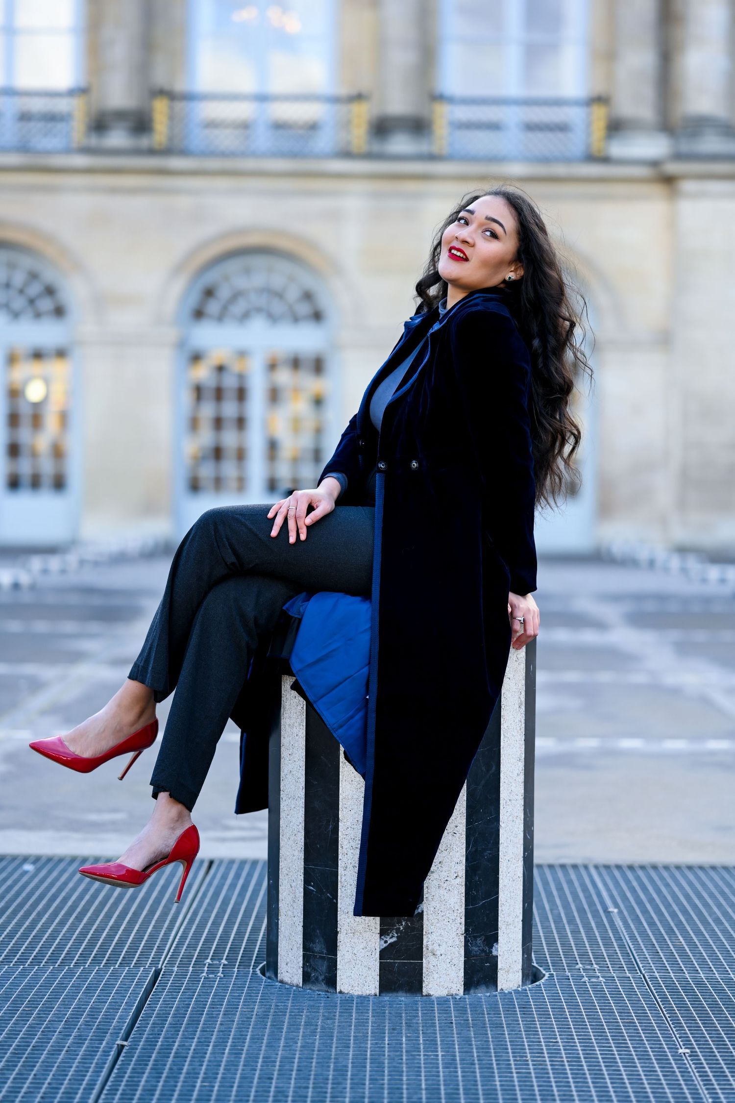 Fashion portrait of woman in black coat sitting on striped column with red heels.