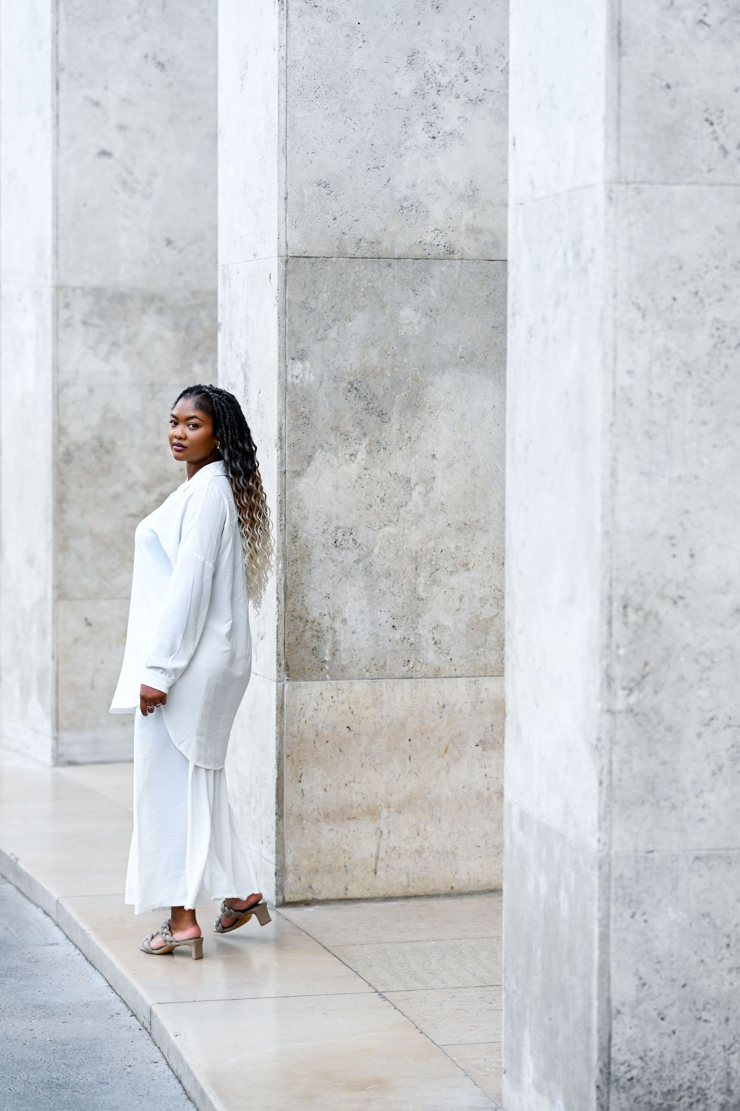 Figure in white outfit poses between modern concrete columns in minimalist setting.