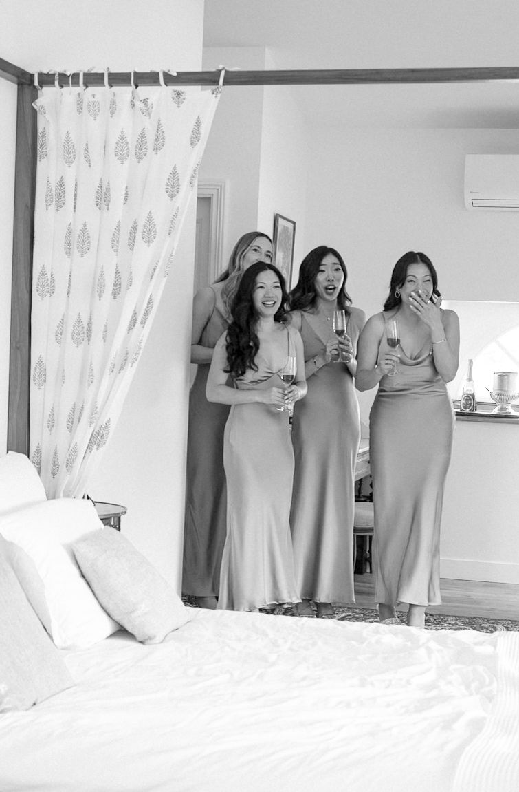 Four bridesmaids in elegant satin gowns react with joy and surprise, holding champagne glasses in a bright bedroom.