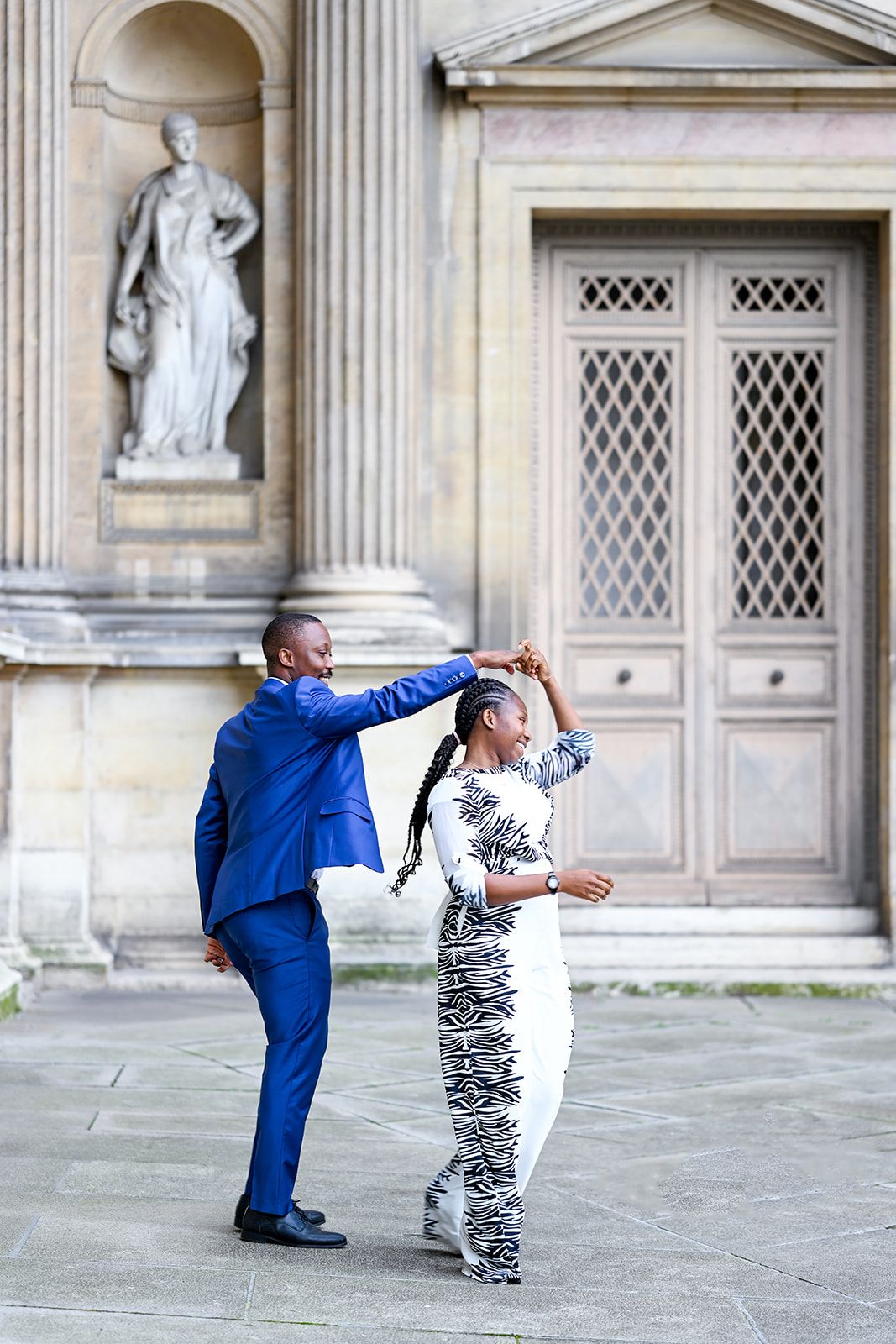 Two people strike dance poses in front of an ornate building with columns and statues.