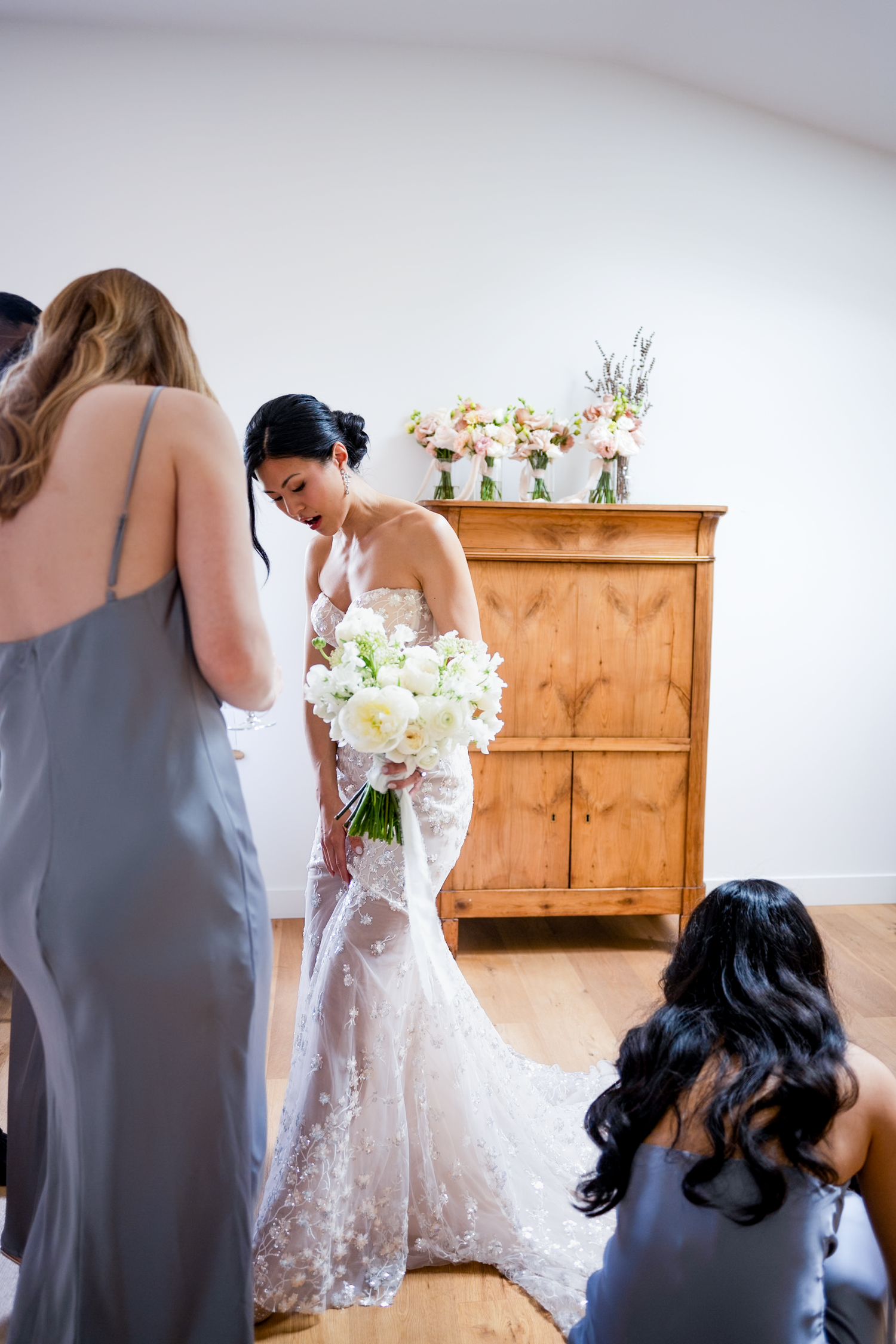 Bride in lace wedding gown holds white bouquet while bridesmaids in gray dresses assist her before the ceremony.