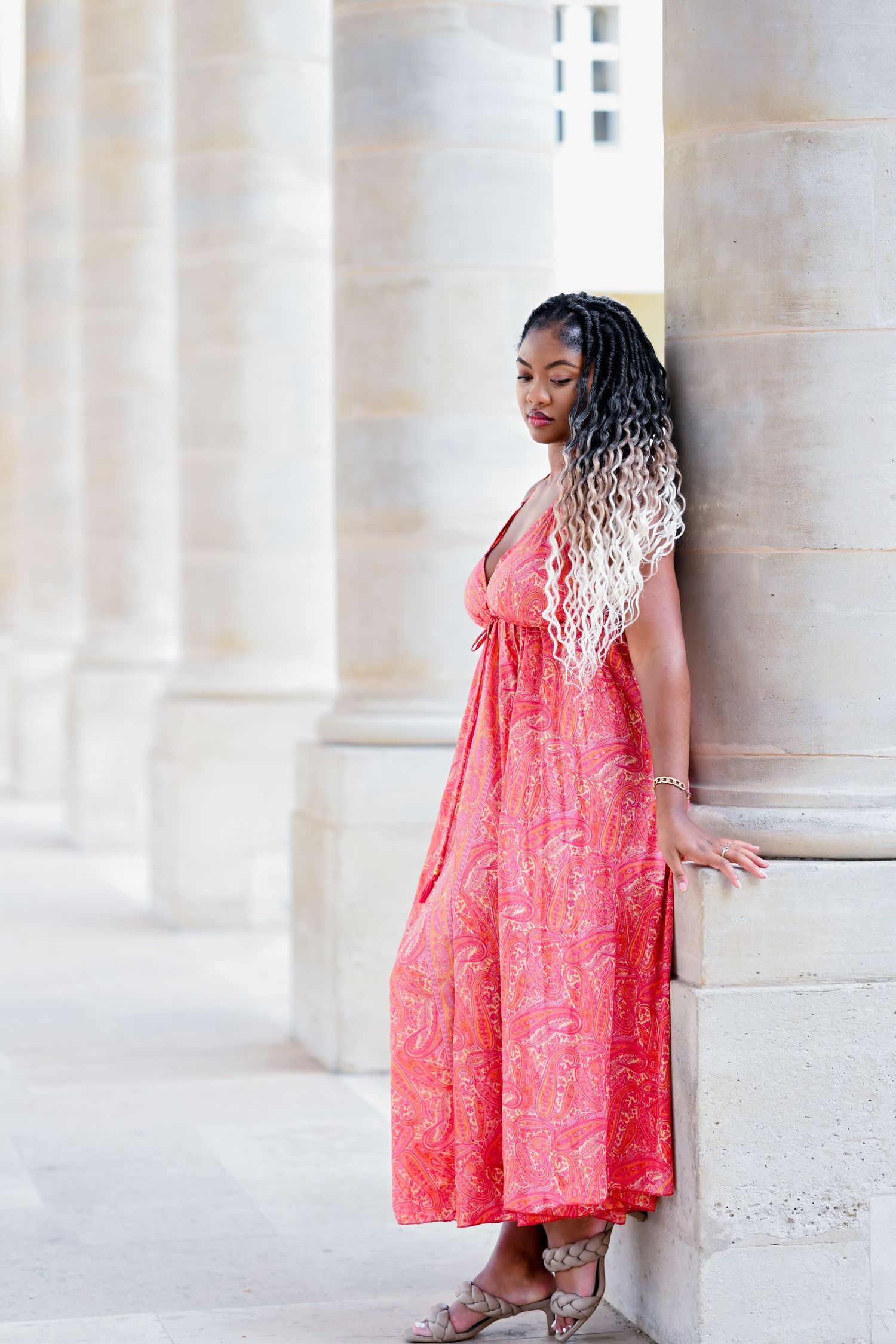Long braided hair cascades down as a coral dress contrasts against white stone columns.