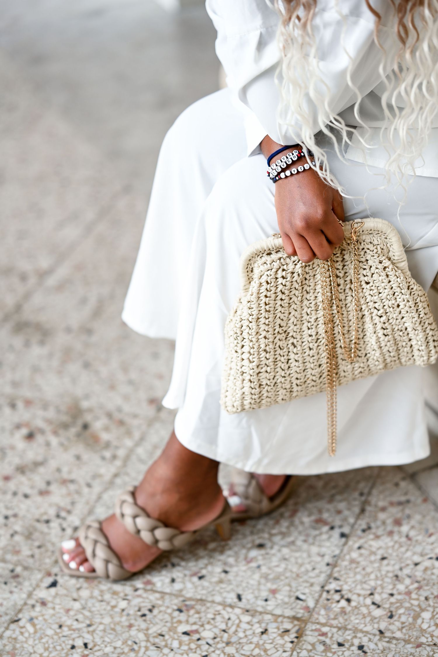 Close-up of woven straw clutch and sandals with white dress on stone floor.