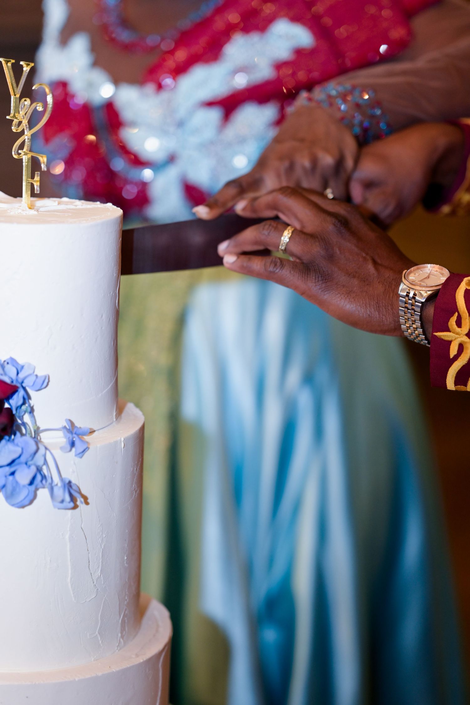 Close-up of hands cutting an elegant white wedding cake adorned with blue flowers.