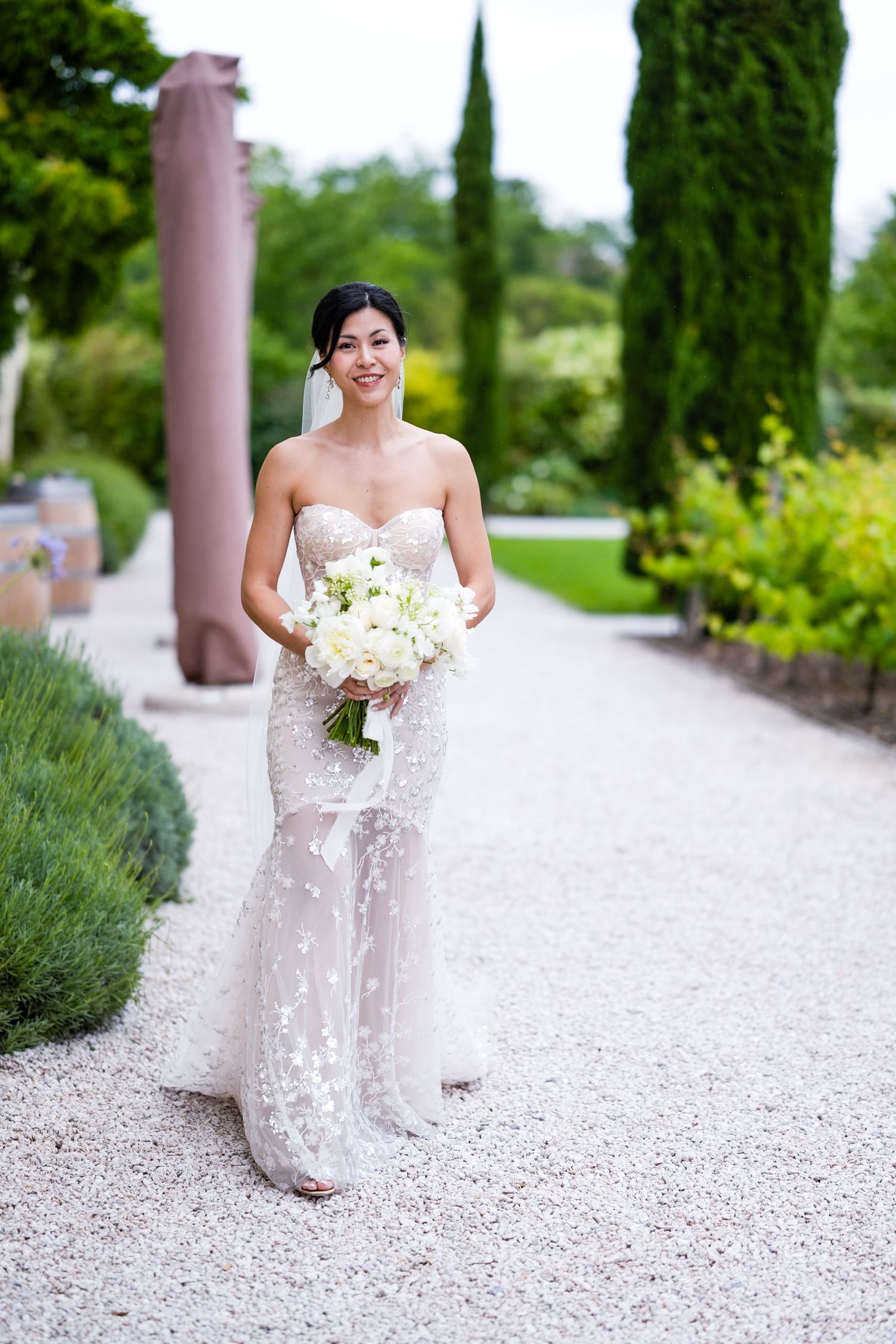 Elegant bride in lace gown holds white bouquet while standing on garden path.