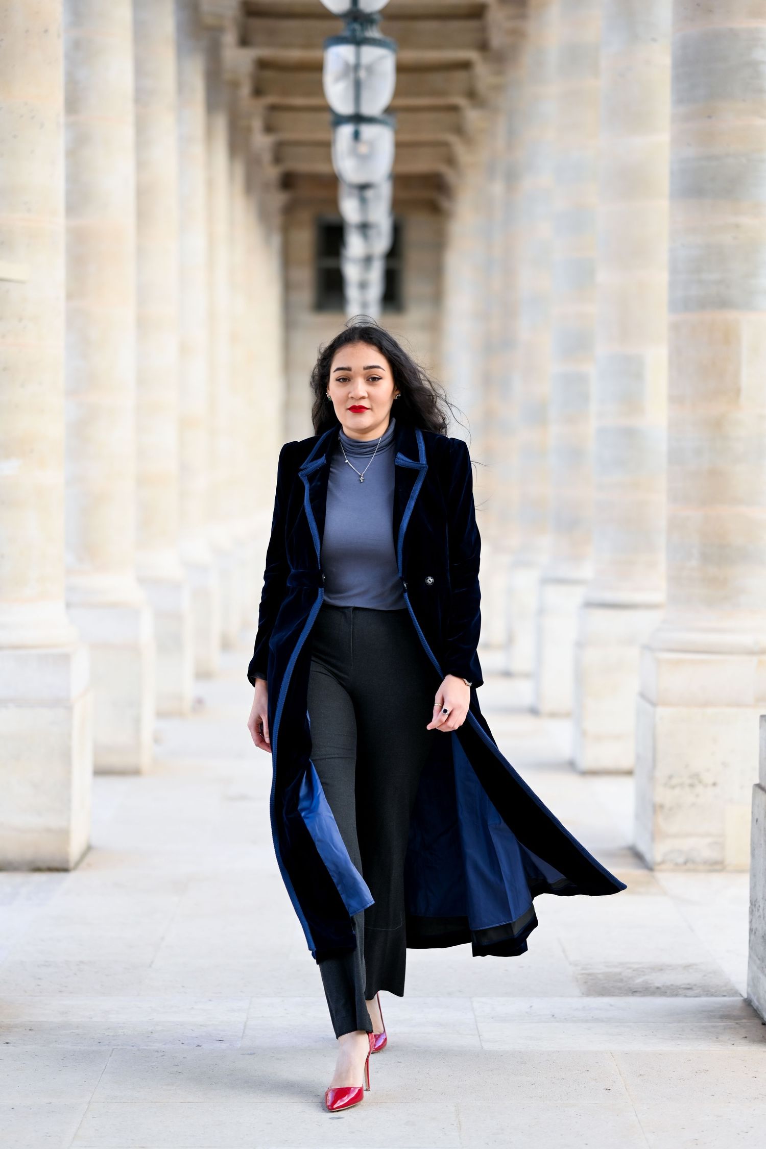 Elegant fashion photo of a long navy coat and red heels in a colonnade hallway with dramatic lighting.