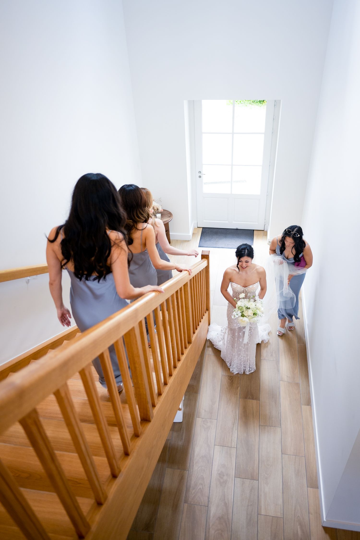 Bride in white gown holds bouquet as bridesmaids in gray dresses descend a wooden staircase toward a bright front door.