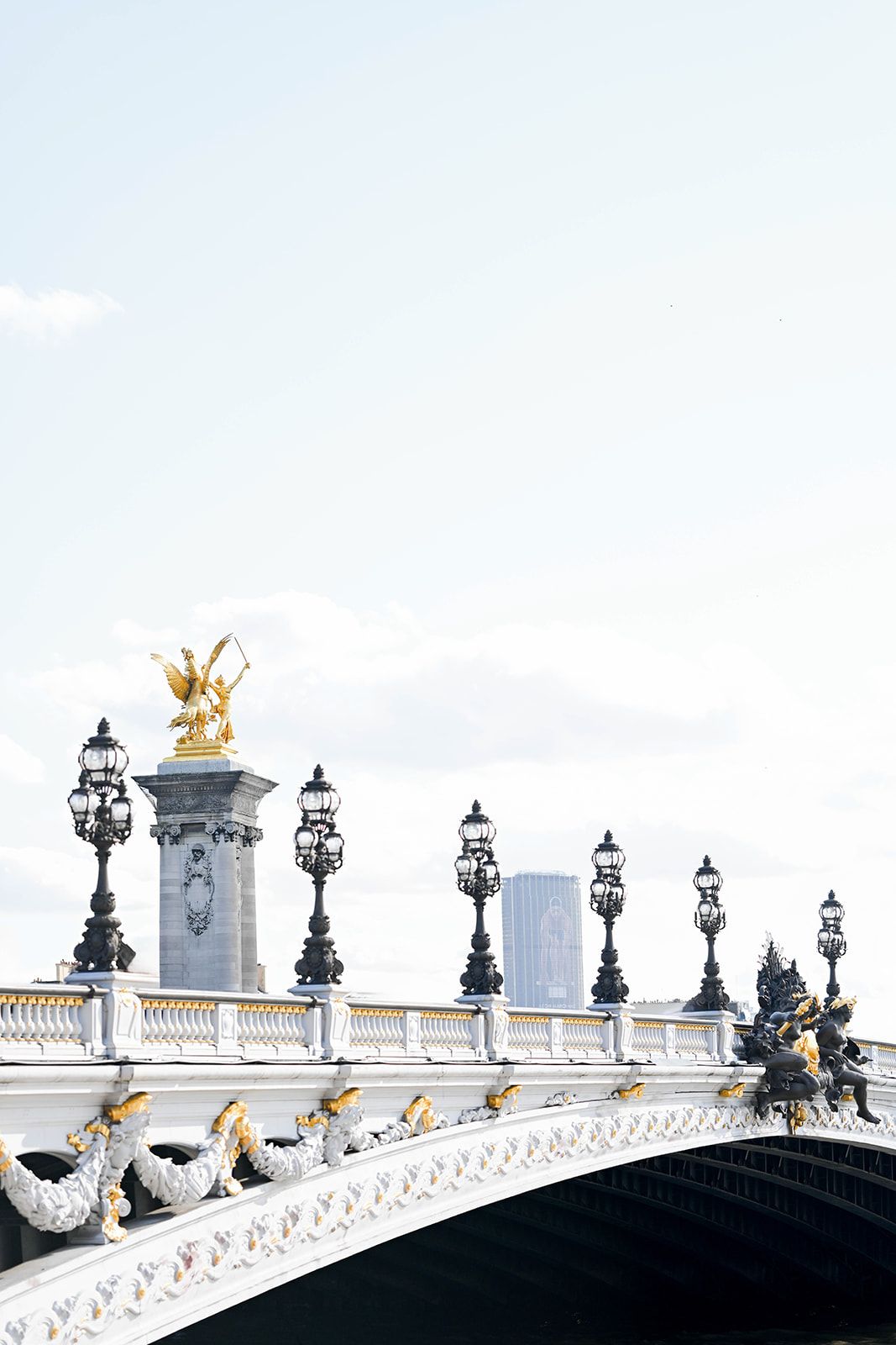 Elegant white bridge with golden statues and ornate lampposts against pale blue sky.