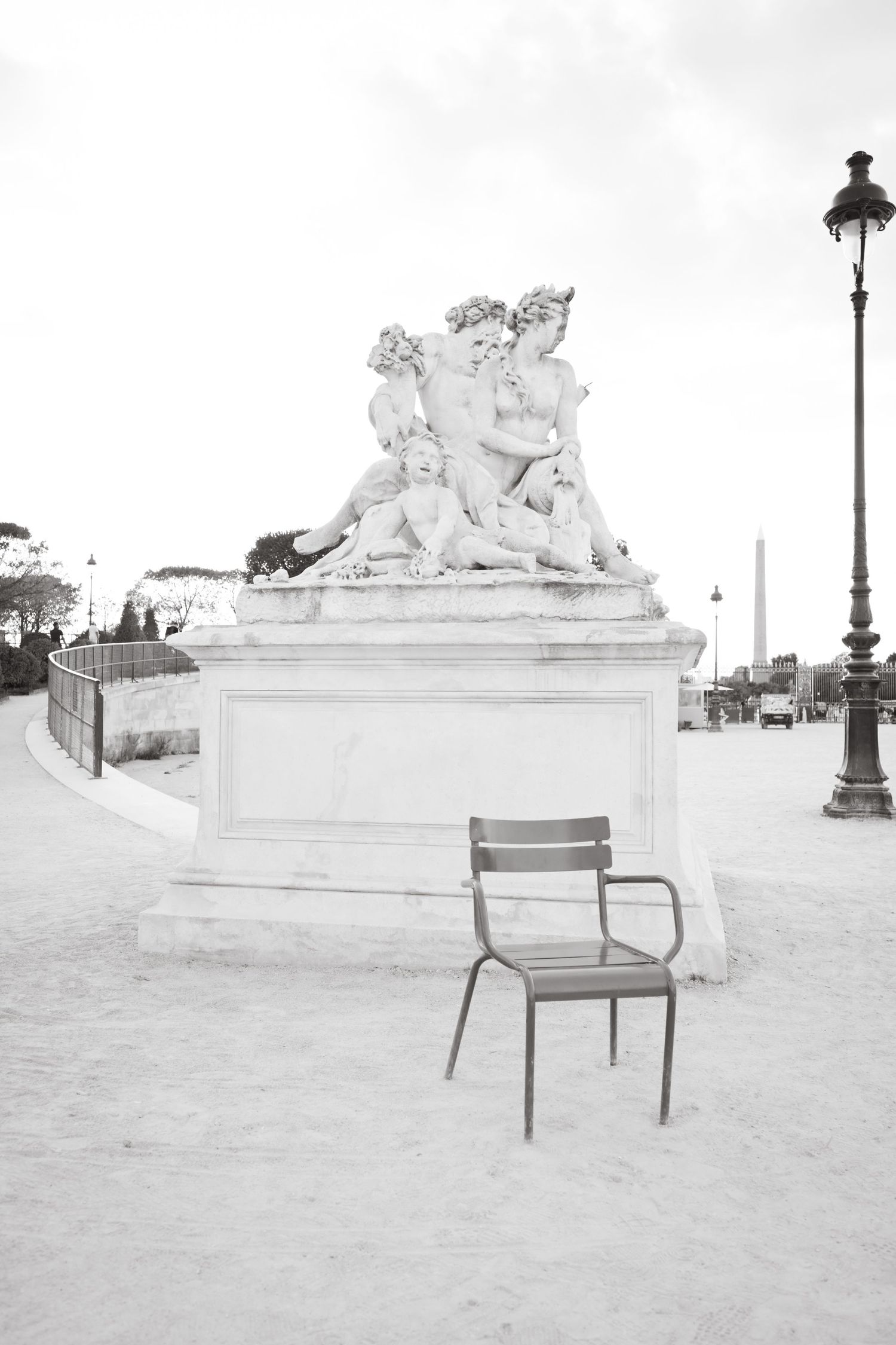 An empty chair sits in front of a classical stone sculpture in a Parisian garden.
