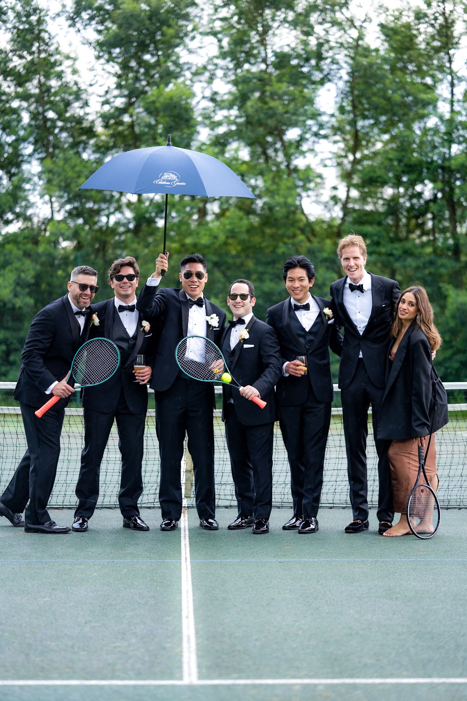 Groomsmen pose playfully with tennis rackets on court in formal tuxedos.