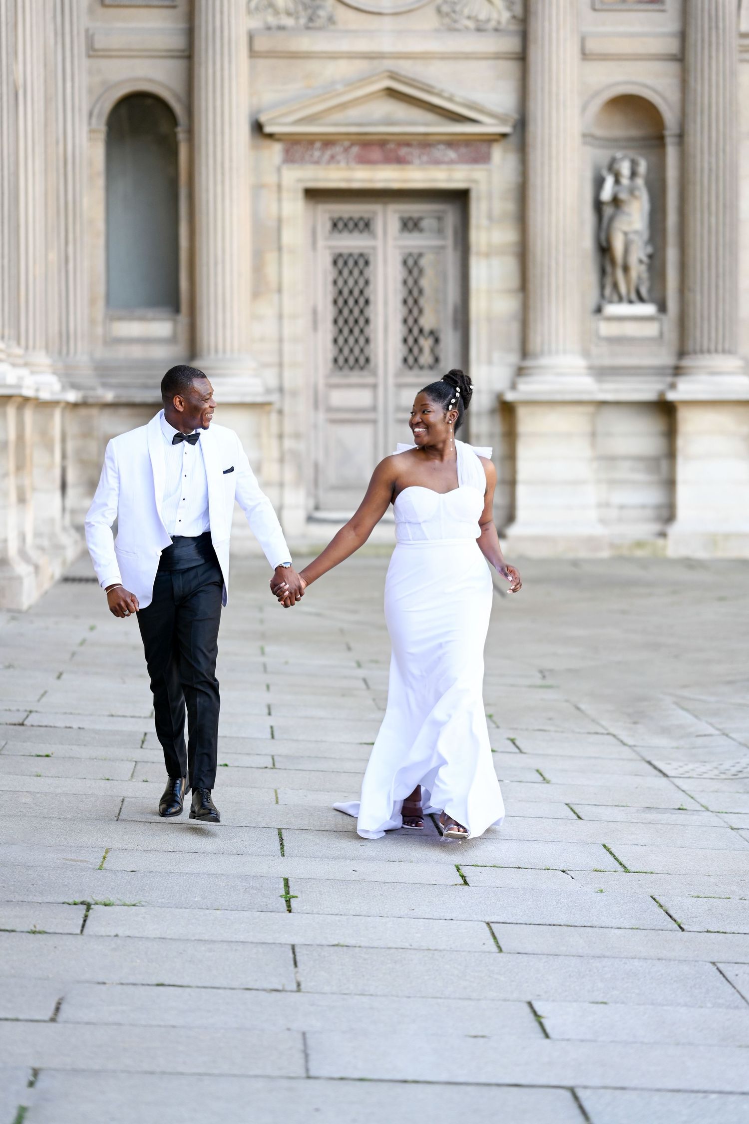 Wedding couple dances playfully on stone courtyard in front of historic building facade.