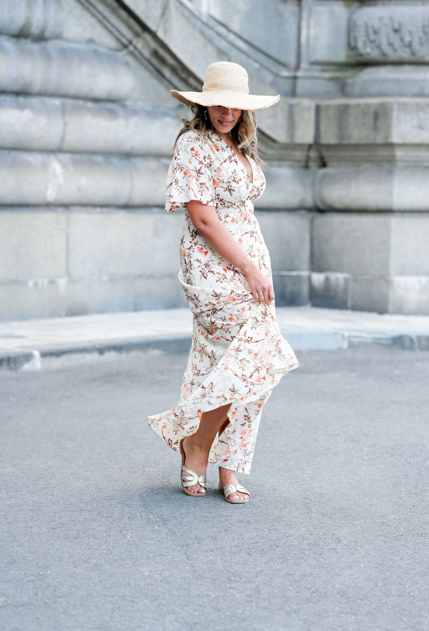 A person in a flowing floral maxi dress and wide-brimmed hat twirls on a city street against a stone building.