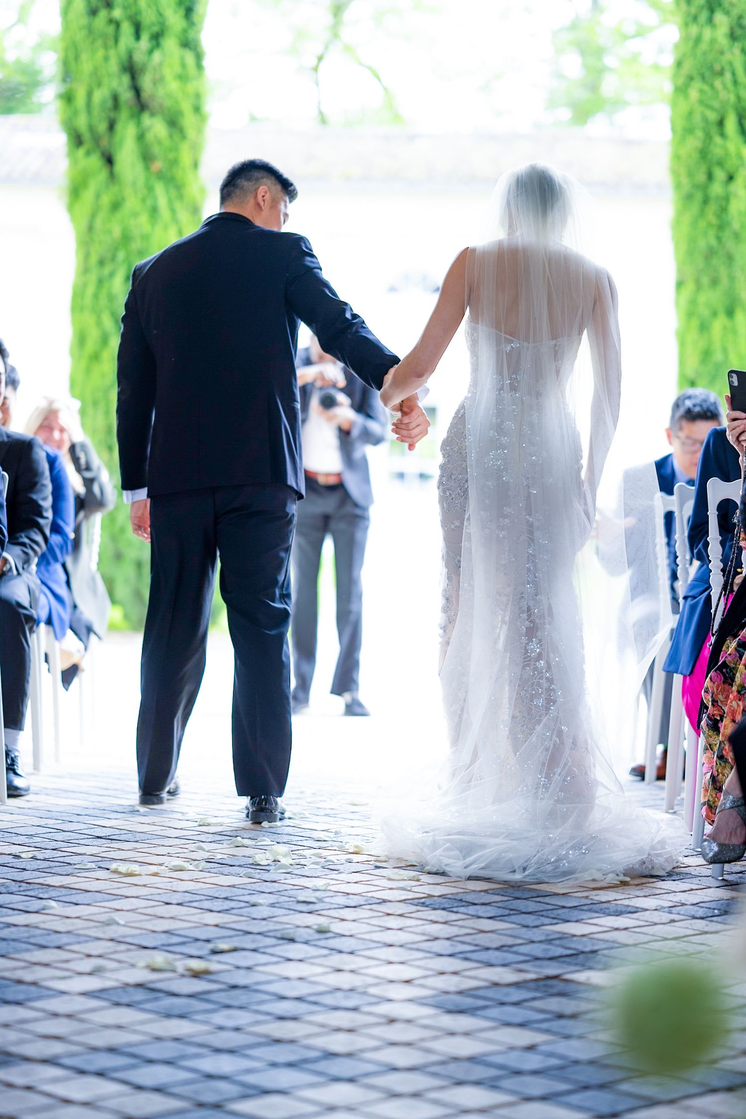Newlyweds walk down the aisle together while guests watch from both sides under a covered walkway.